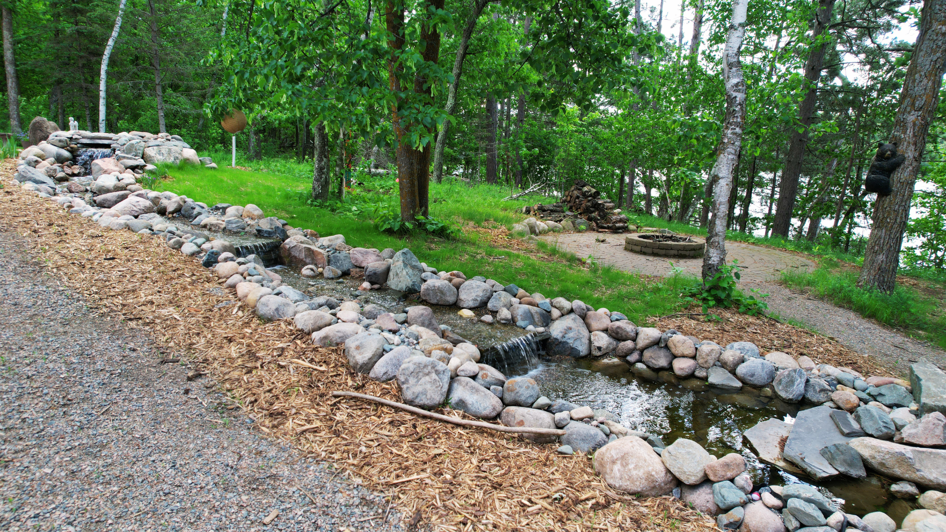 A backyard with a small stream bordered by rocks, surrounded by green grass and trees, with a fire pit and stacked firewood in the background.