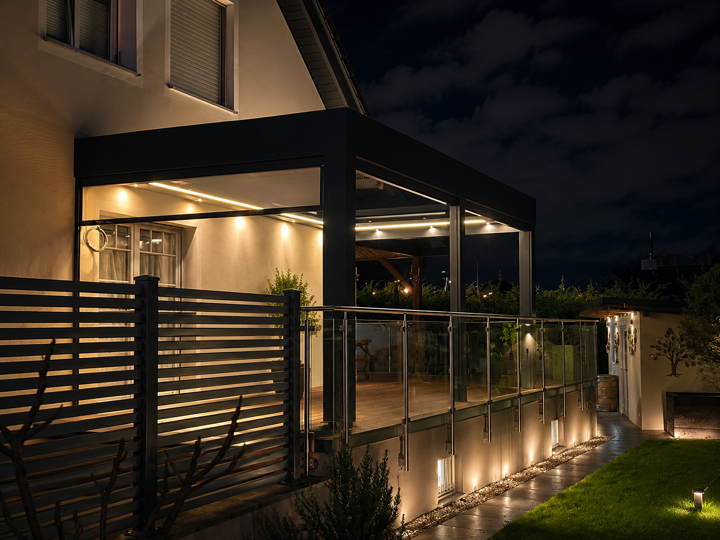 Nighttime view of a modern house's outdoor patio with glass railing, warm lighting, and a covered section, surrounded by a lawn and decorative elements.