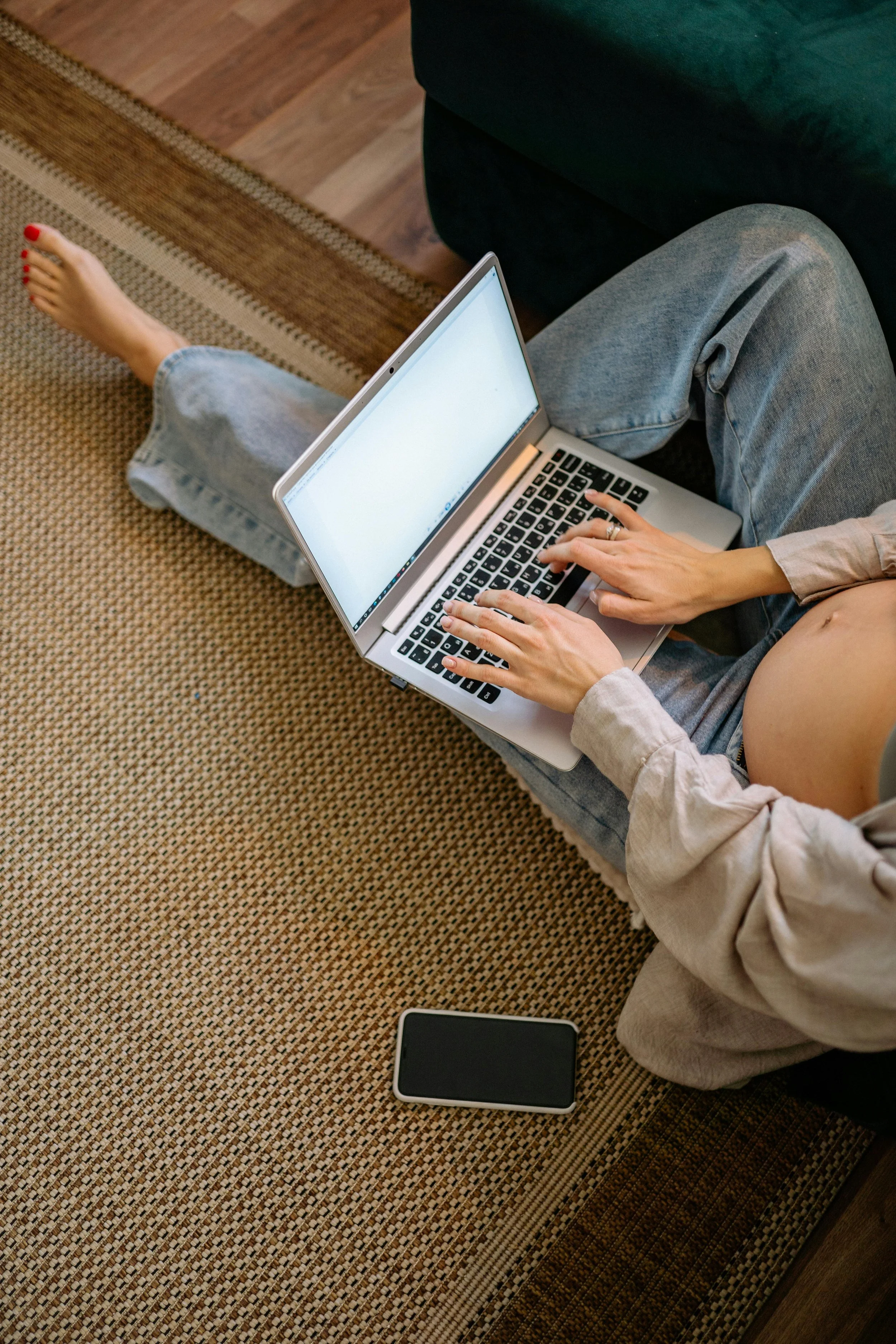 A person sitting on a rug with crossed legs, using a laptop on their lap and a smartphone resting on the rug nearby.