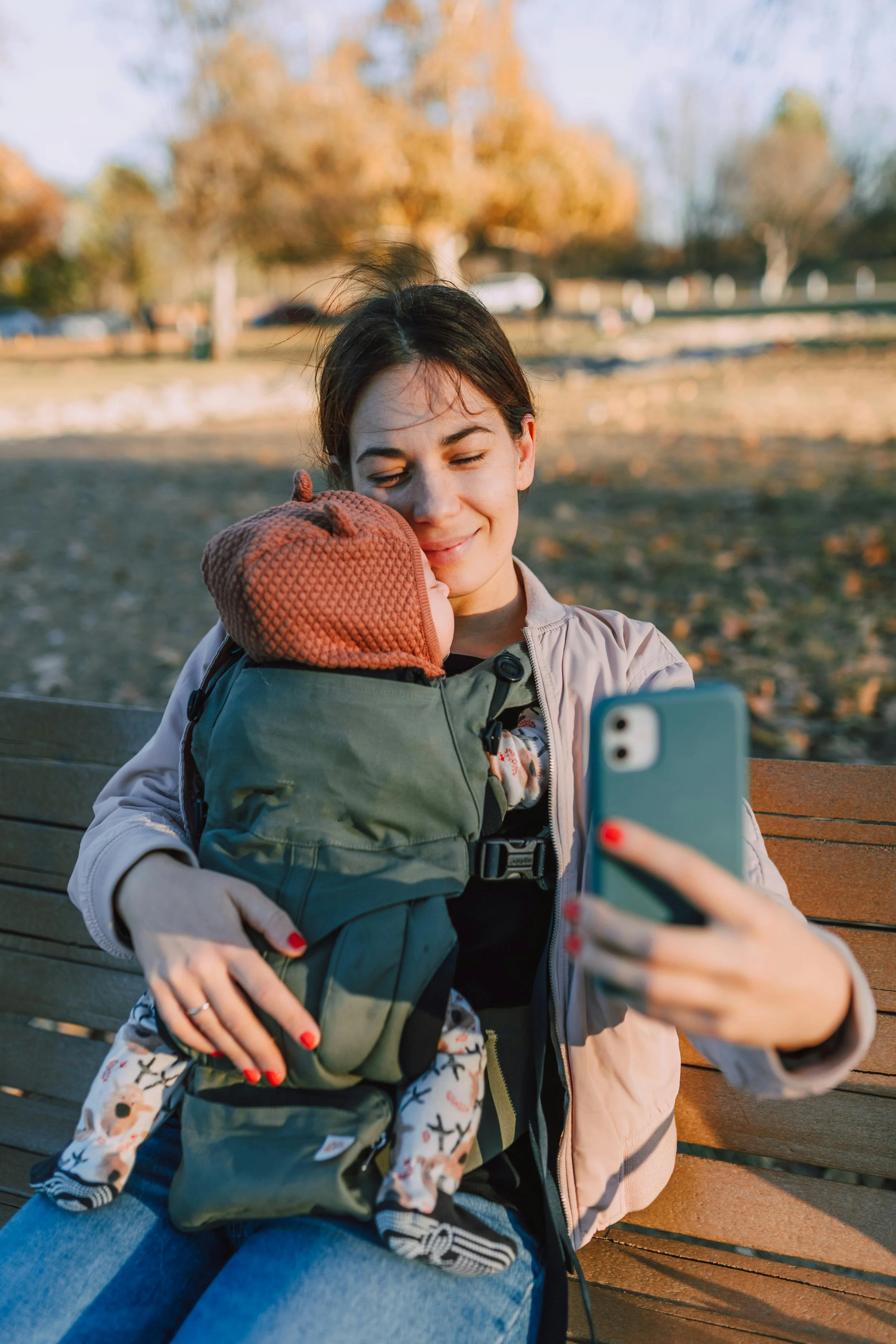 A woman sitting on a park bench, holding a baby in a front carrier, taking a selfie with her smartphone during daytime.