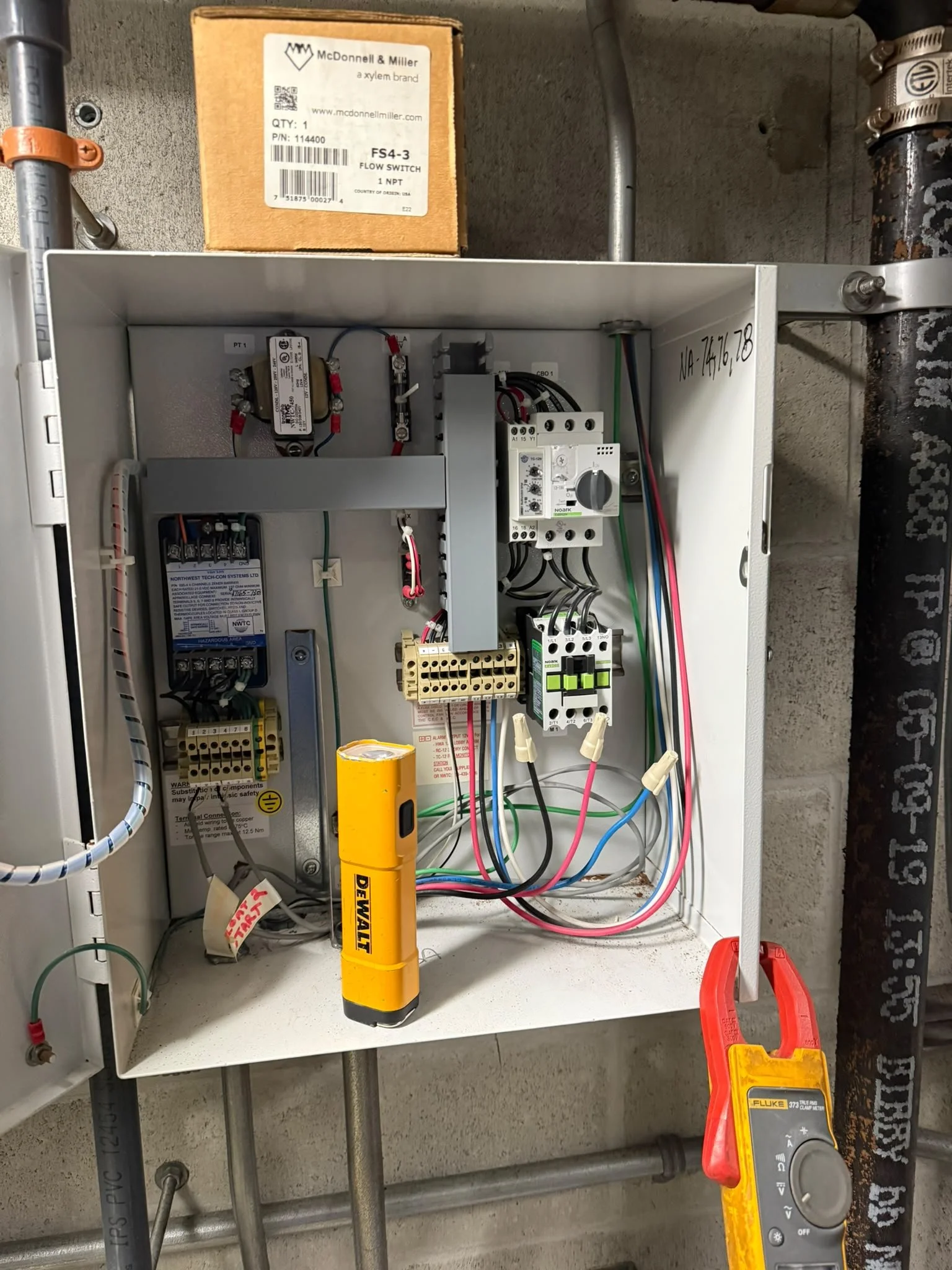 Open electrical control panel with wires, circuit breakers, and components, a multimeter, and a yellow DeWalt tool on a white surface.