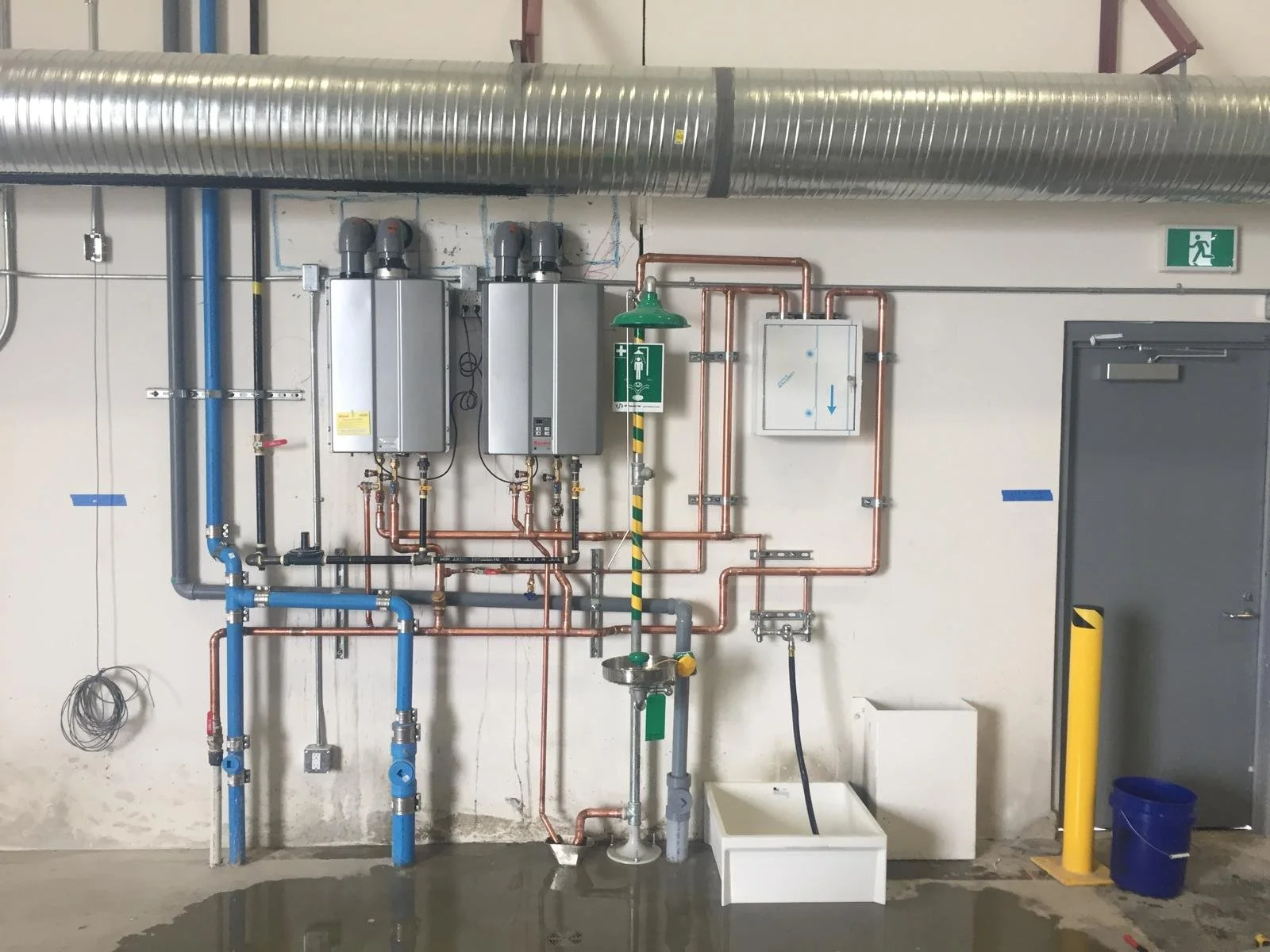 Industrial piping and equipment on a beige wall, including metal, copper, and plastic pipes, control boxes, a drain, a sink, and safety signs.