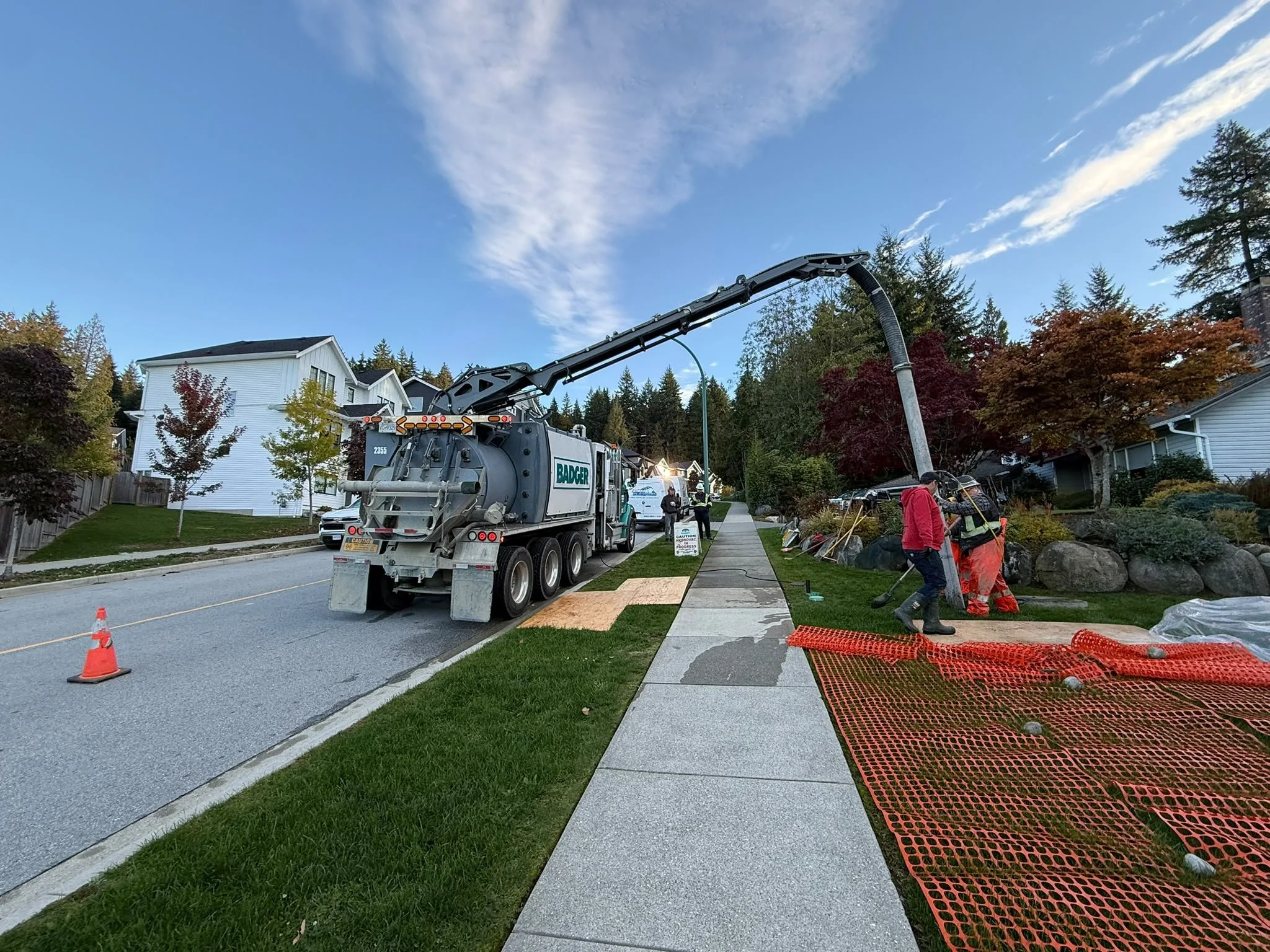 A street scene with a large vacuum truck extending its hose into a house for cleaning or repairs, with workers nearby, traffic cones, and orange safety fencing on the sidewalk, under a partly cloudy sky.