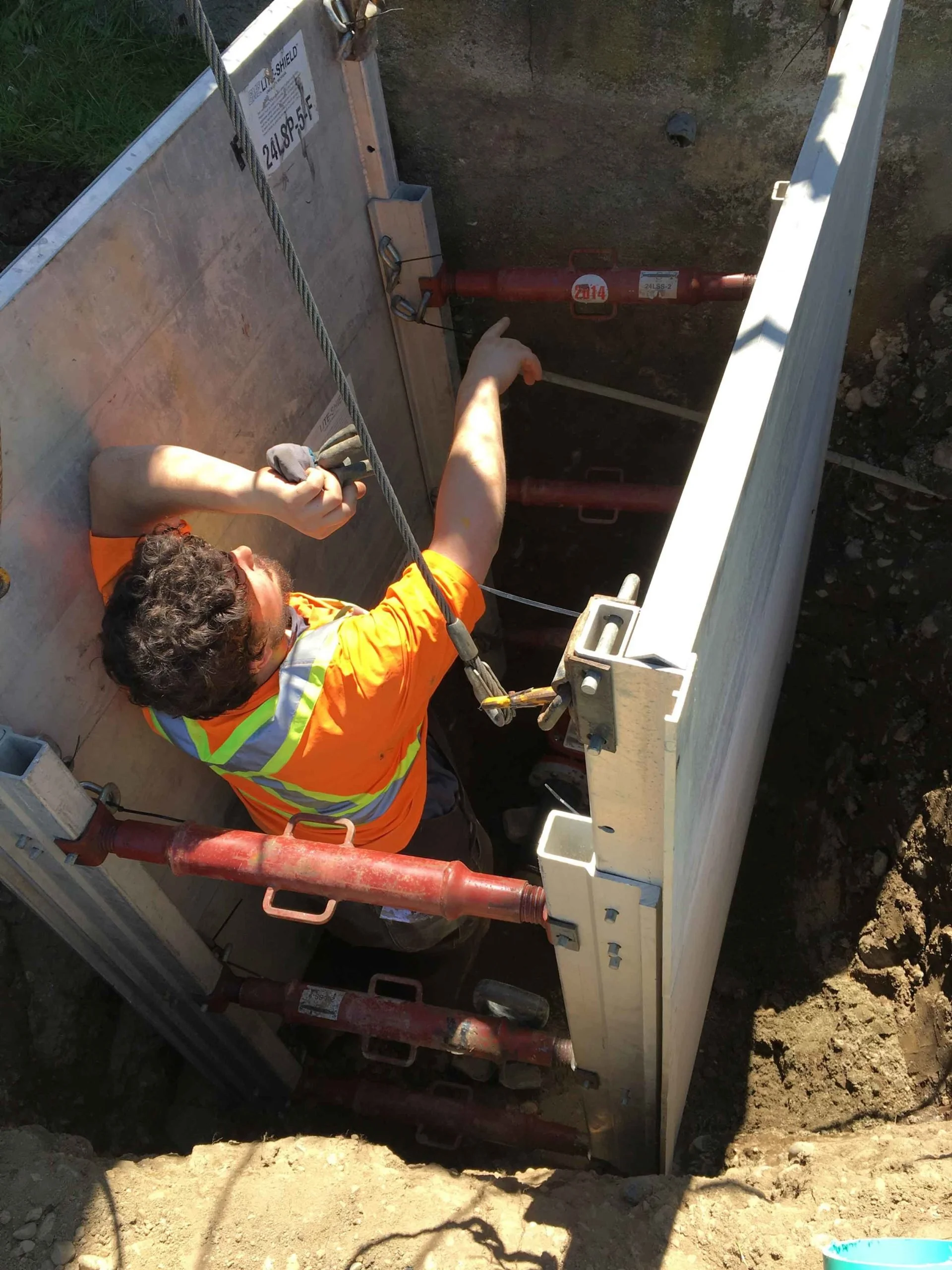 Worker in orange safety vest and gloves operates machinery inside an excavation site, with dirt and equipment visible around.