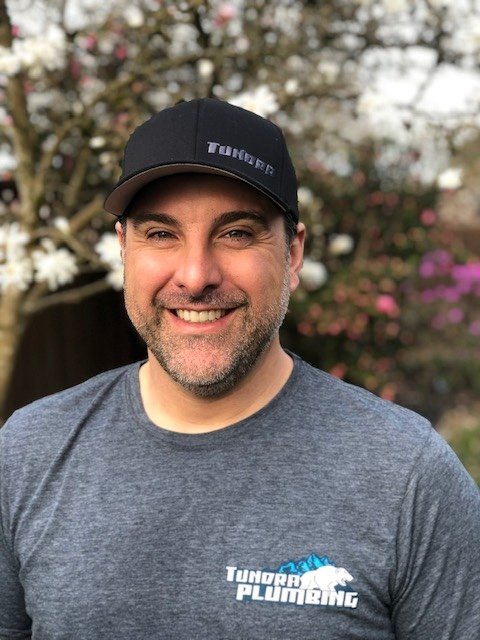 A smiling man wearing a black cap with 'TUNNER' written on it, and a gray T-shirt with 'TUNNER PLUMBING' logo, standing outdoors with blooming bushes in the background.