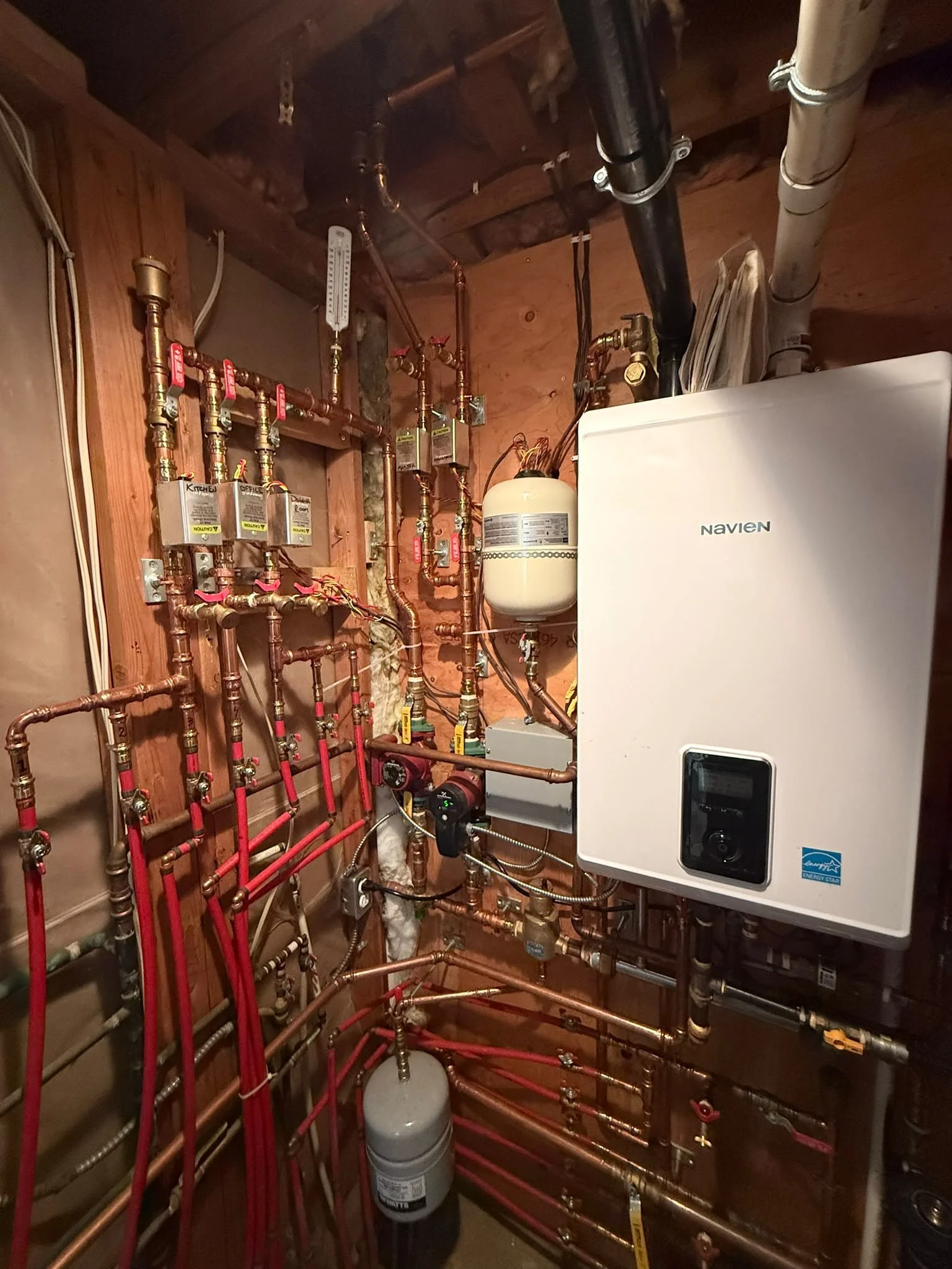 A utility room containing a Navien tankless water heater, copper piping, a expansion tank, and a digital thermostat for the heating system. The room has unfinished wood walls and ceiling.