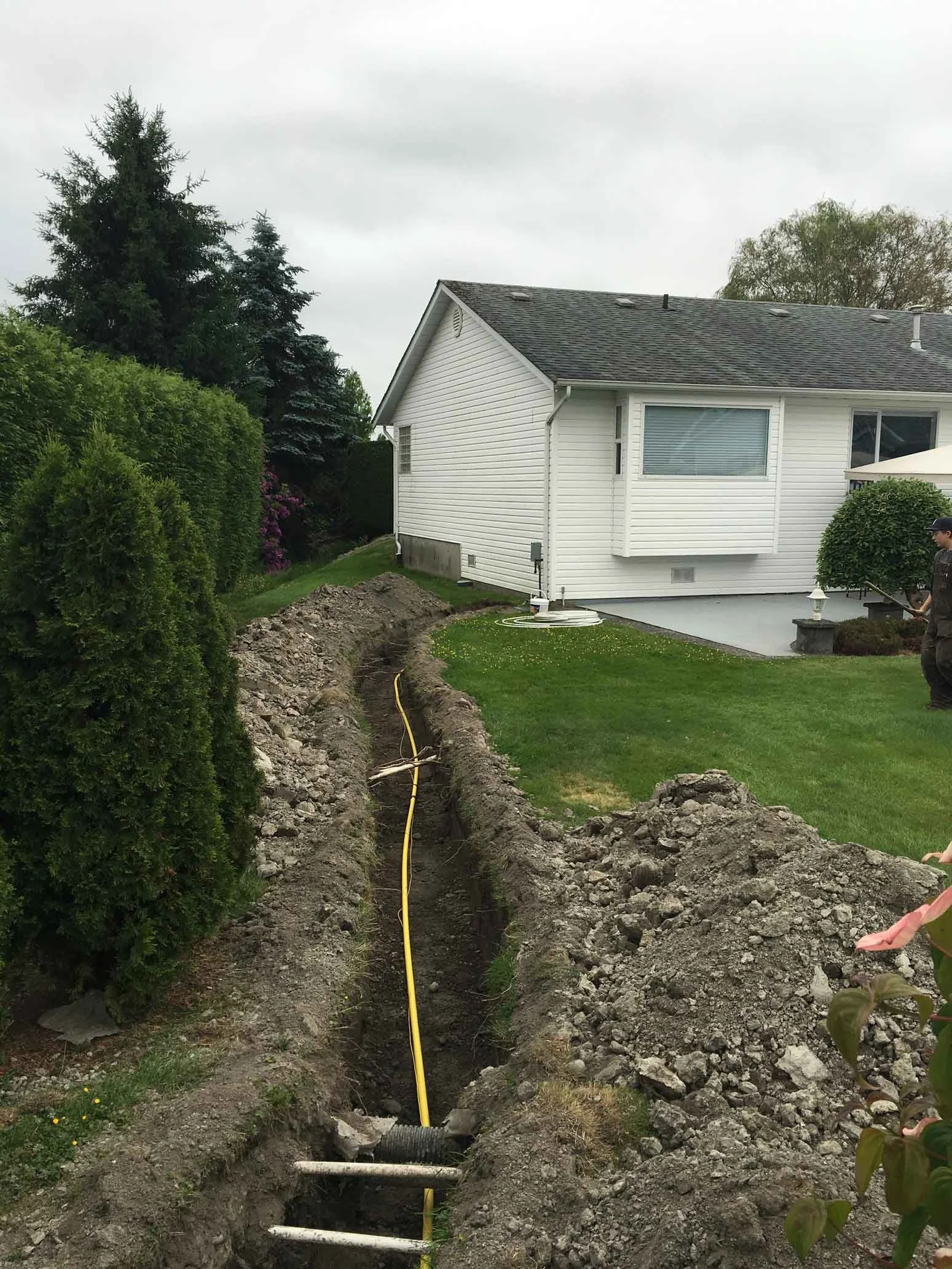A backyard with a trench dug for underground utility work, a yellow hose running through it, and a white house with a window, bushes, and a person wearing a cap walking near the trench.