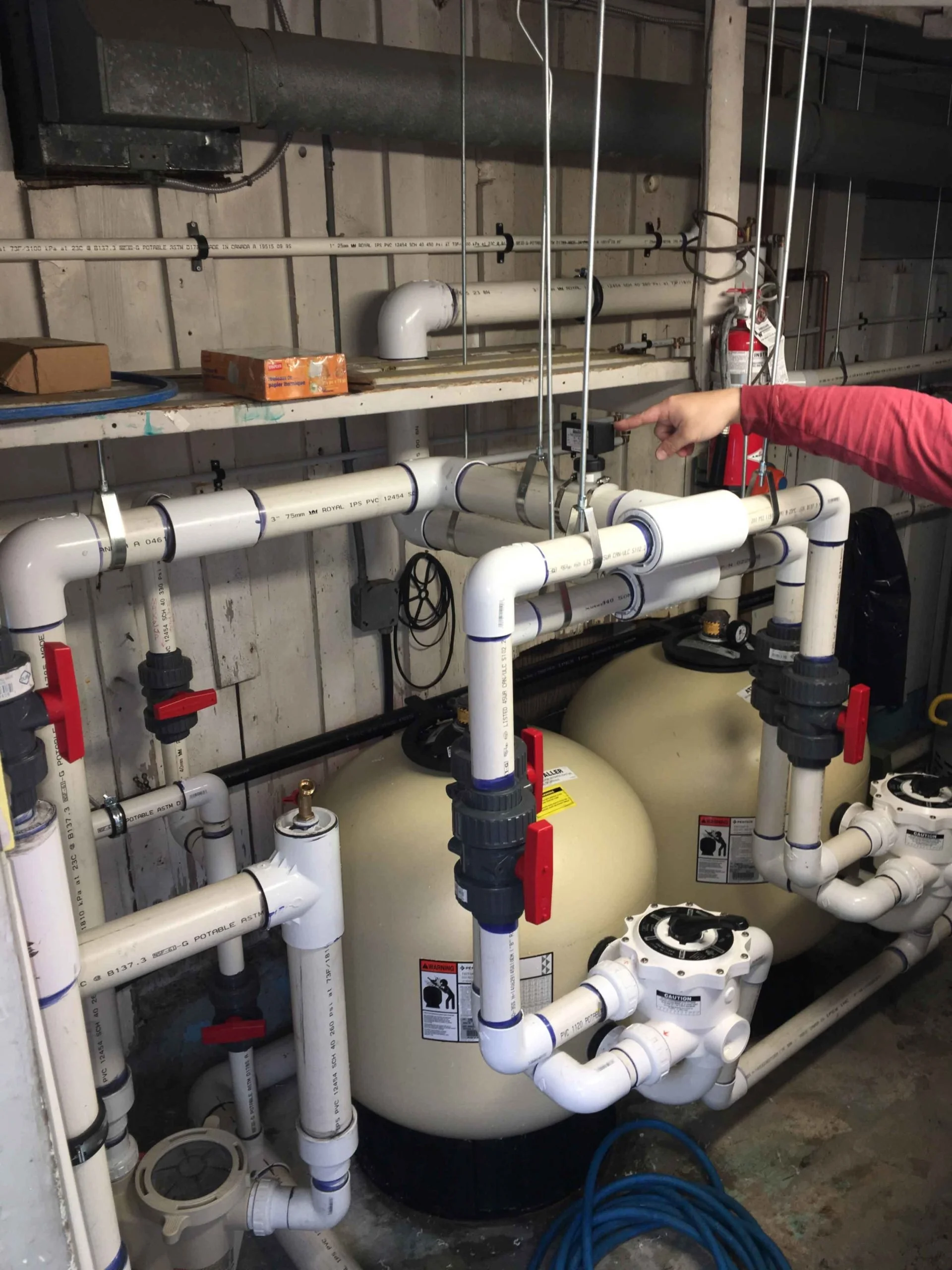 A plumbing system with white PVC pipes, valves, and a large round tank, with a person pointing at the setup. The pipes are arranged against a concrete wall and are connected with various fittings and control mechanisms, including a fire extinguisher 