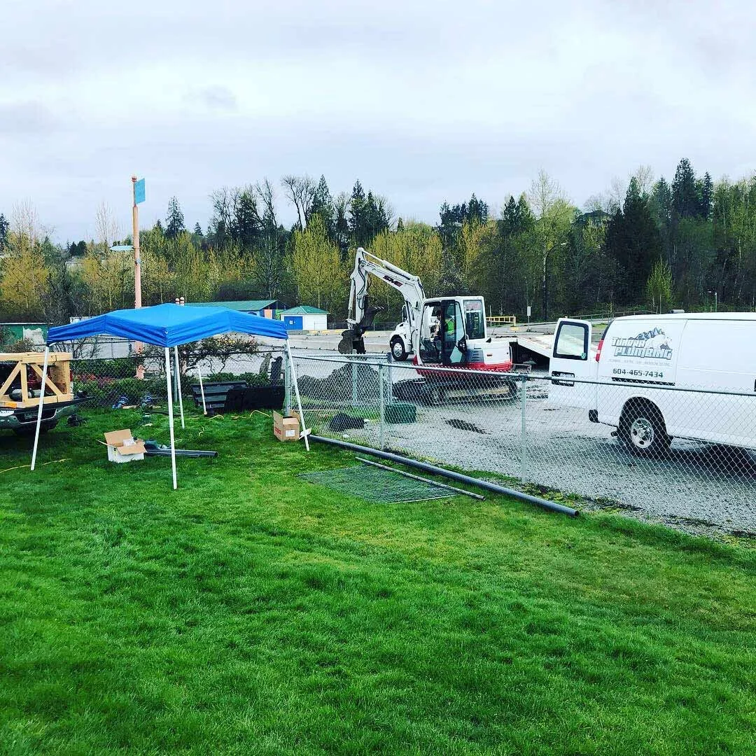 Construction site with a blue canopy tent, a small white excavator, and a white utility van behind a chain-link fence, set on a grassy area.