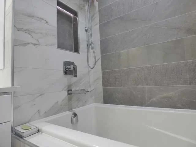 Bathroom with a bathtub, a handheld showerhead, a small window, and gray tiled walls.