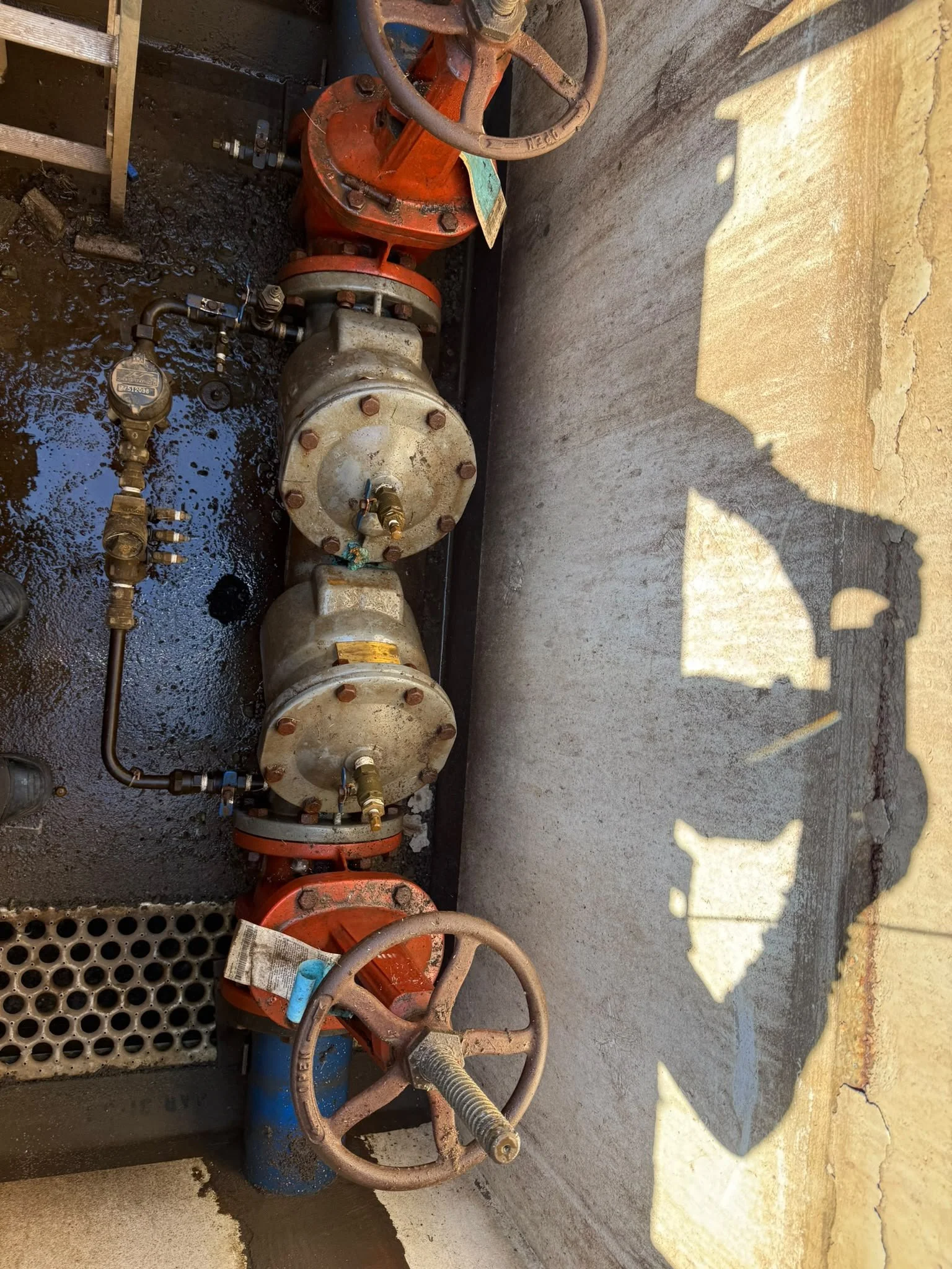 Close-up of industrial piping system with valves, pipes, and fittings, some of which are rusty, installed against a concrete wall. The pipes are connected by various fittings and valves, with some dark liquid or residue on the surface.