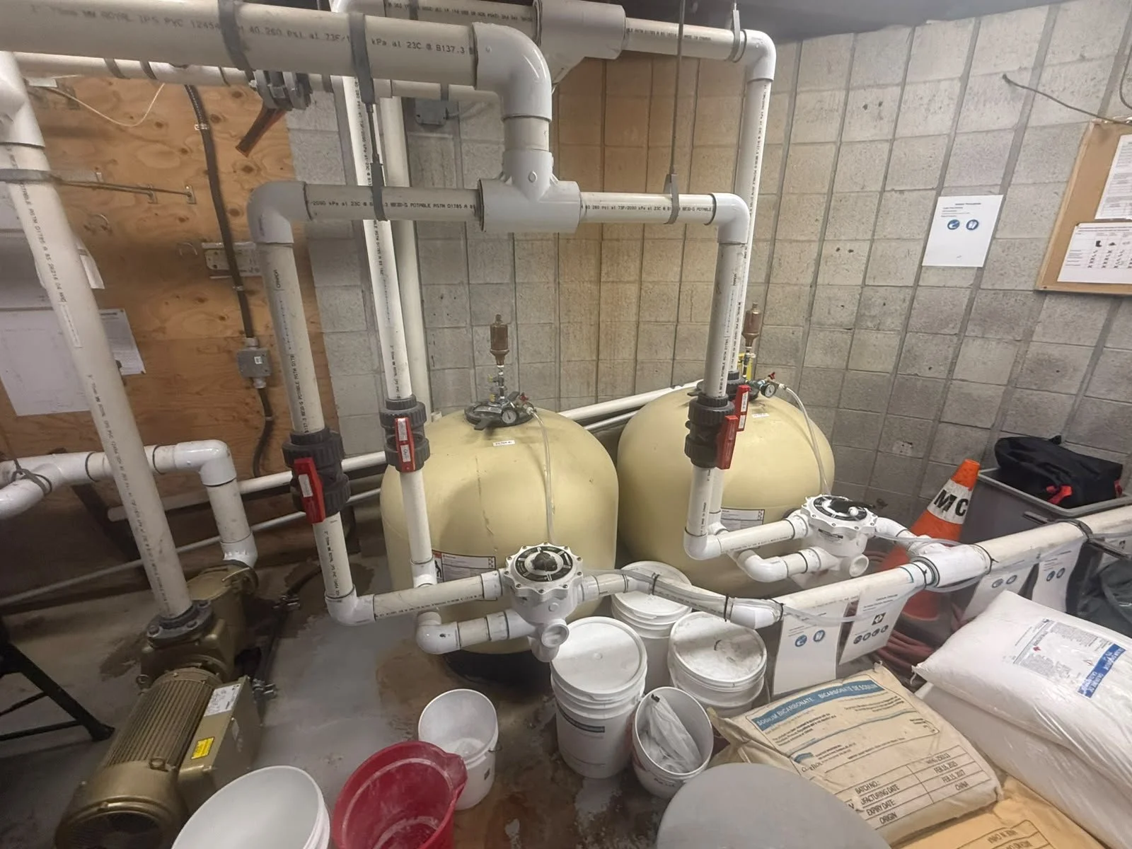 The image shows a mechanical room with plumbing system, including white PVC pipes, valves, and two beige pressure tanks, against a concrete block wall. There are buckets, a toolbox, and construction materials on the floor.