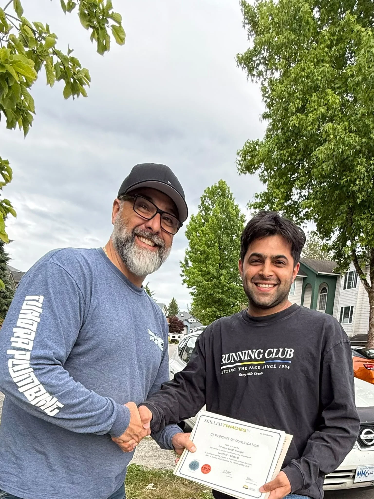 Two men shaking hands, smiling, with one holding a certificate of qualification outdoors in a residential neighborhood.