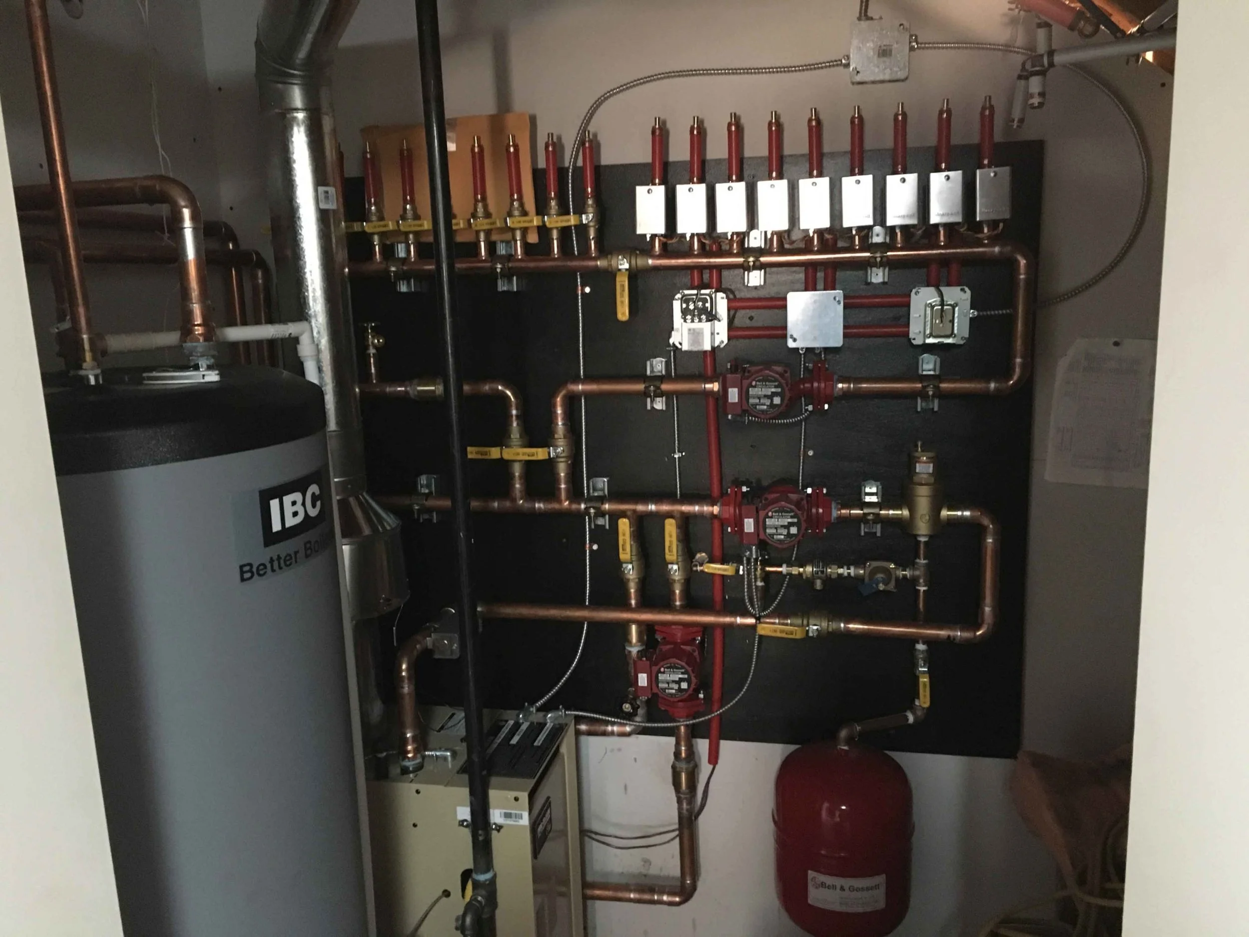 A plumbing system with copper pipes, valves, and a red expansion tank mounted inside a cabinet. There is a water filter labeled 'IBC'.