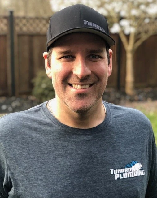 A smiling man wearing a gray Tundra Plumbing T-shirt and a black baseball cap outdoors with a wooden fence and trees in the background.