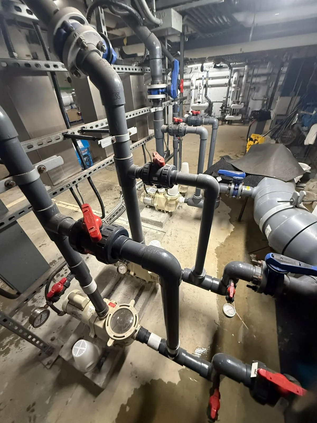 Industrial pipes and valves in a mechanical room, with various black, gray, and red-colored pipe fittings, gauges, and a large duct on the right side.