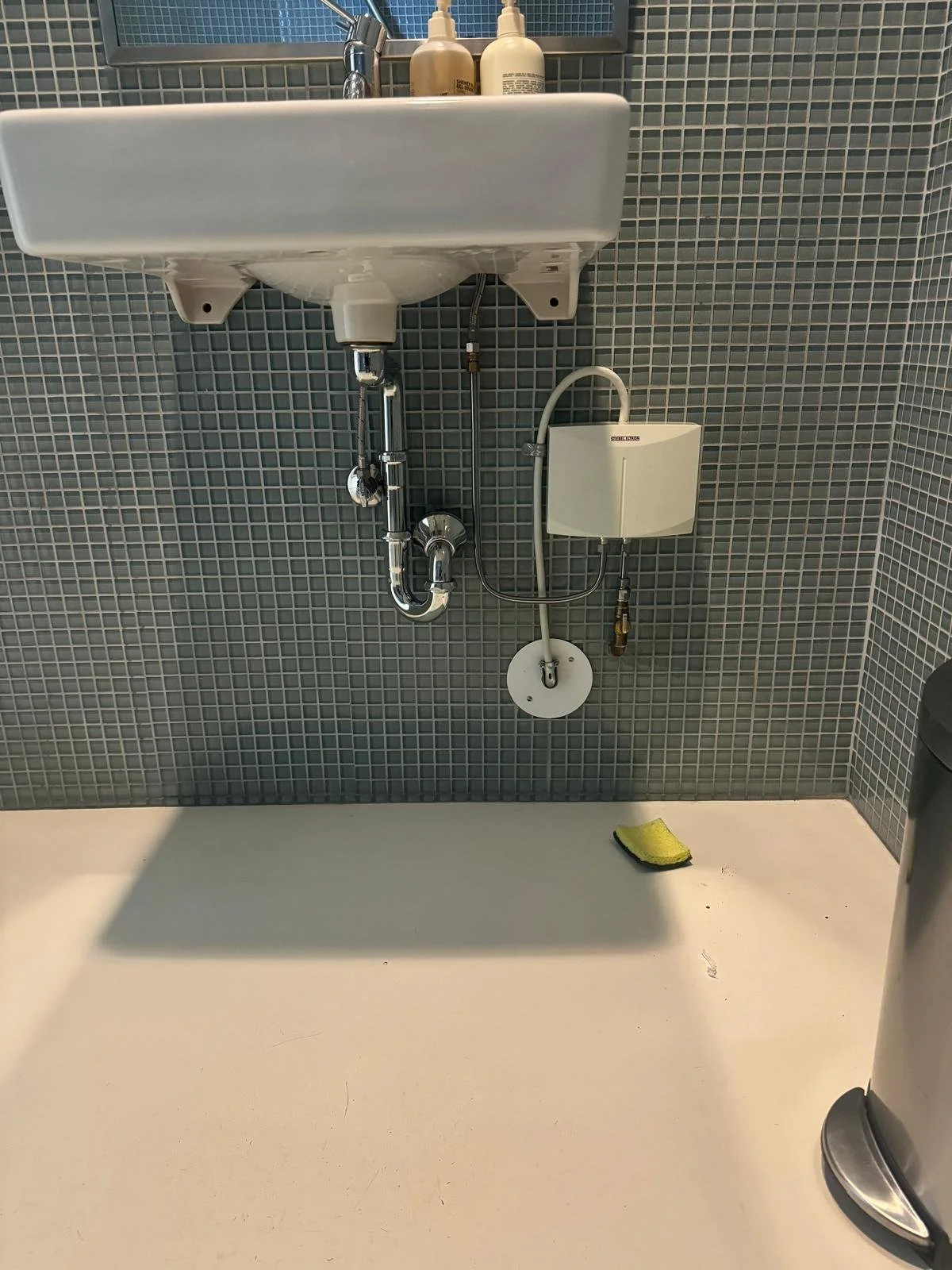 A small bathroom sink mounted on a tiled wall with soap dispensers on top, plumbing exposed beneath, a small white box attached on the wall, a sponge on the white floor below, and part of a trash can on the right.