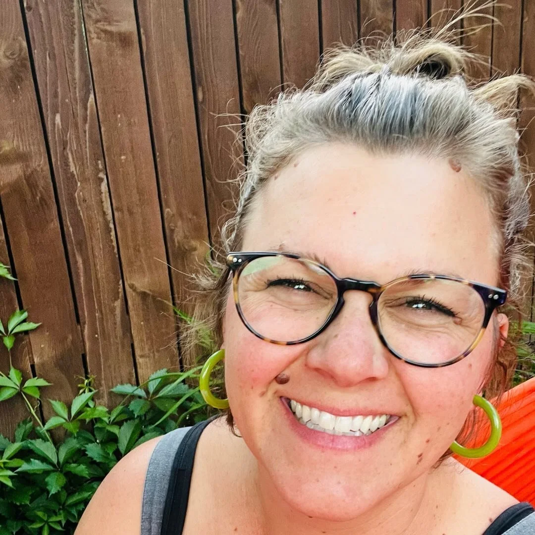 A woman with glasses and large green hoop earrings smiling outdoors in front of a wooden fence and green plants.