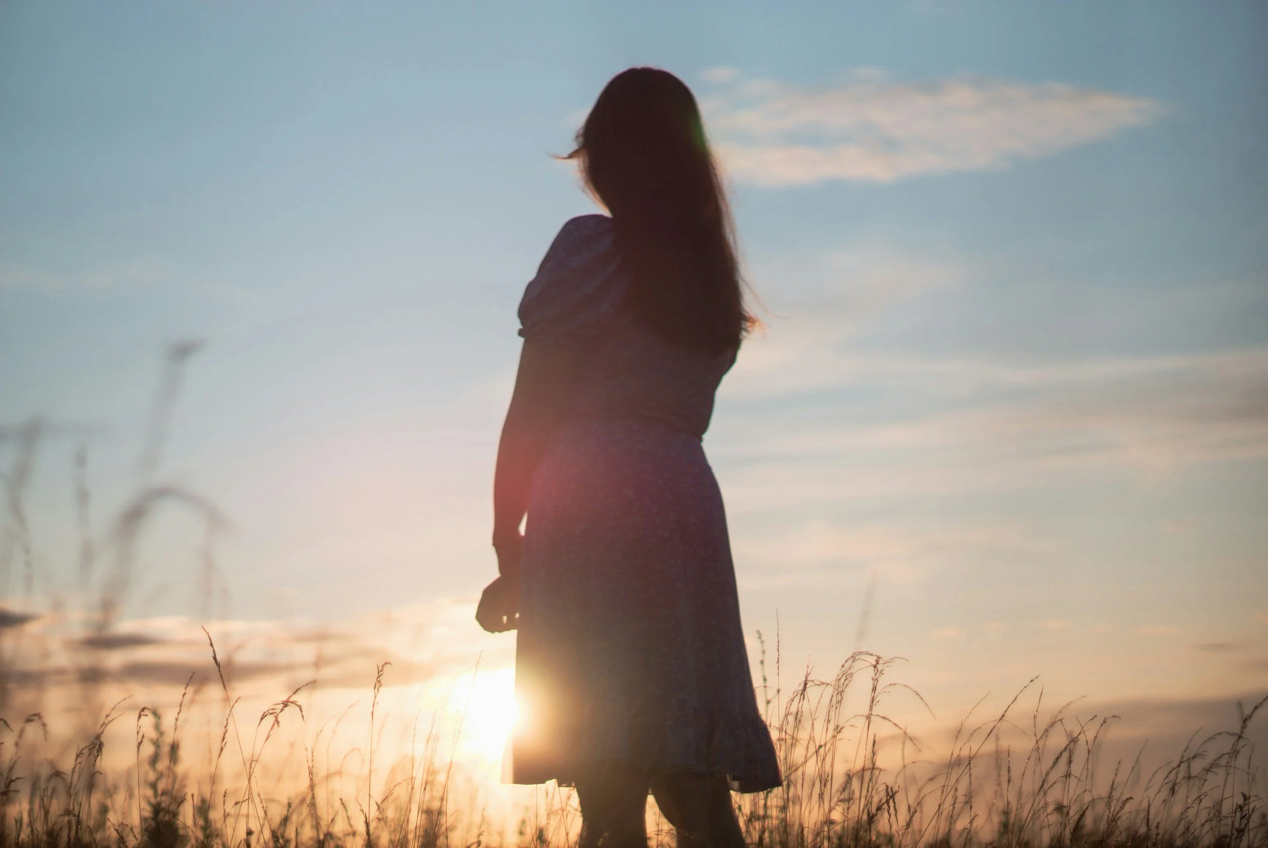 A woman in a long skirt stands in a field during sunset, with her back towards the camera and her hair flowing in the breeze.