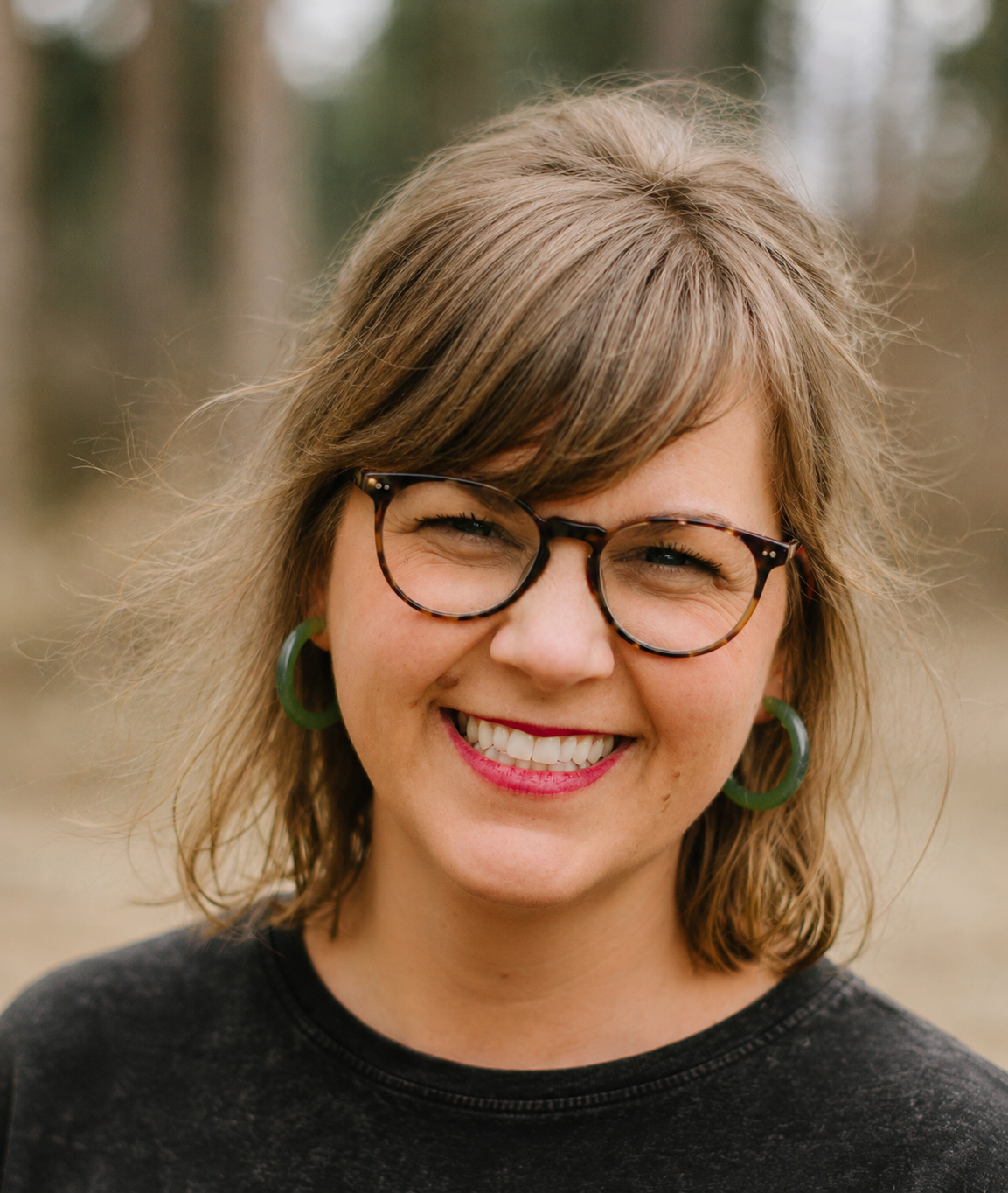 A woman with light brown hair, glasses, and green earrings smiling outdoors.