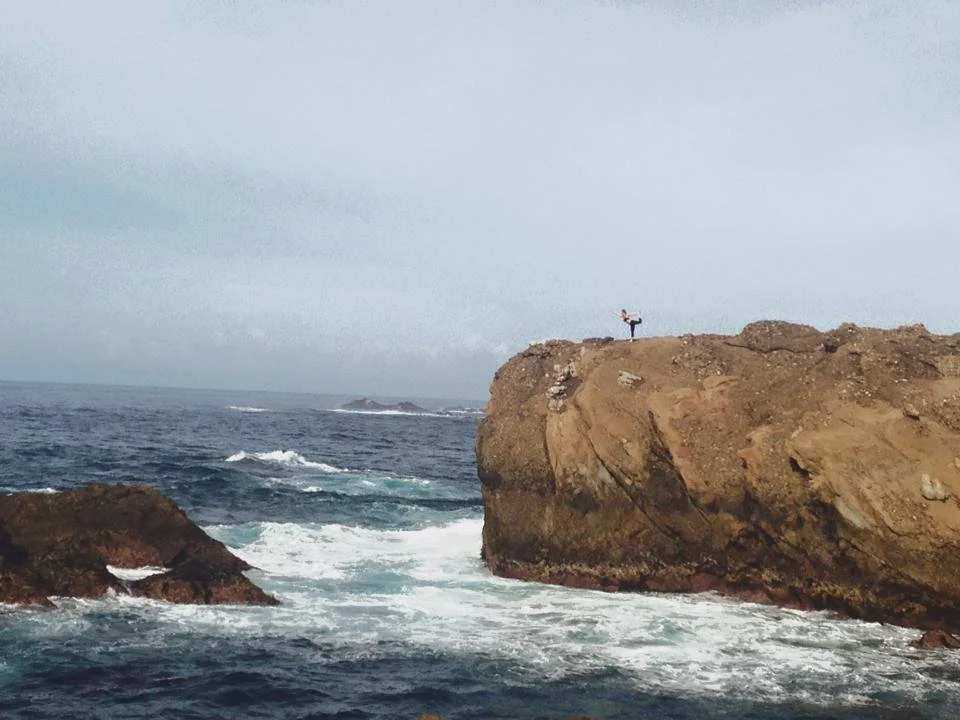 Person standing on a large rocky cliff by the ocean, doing a yoga pose with one arm raised, overlooking the water with waves crashing below.