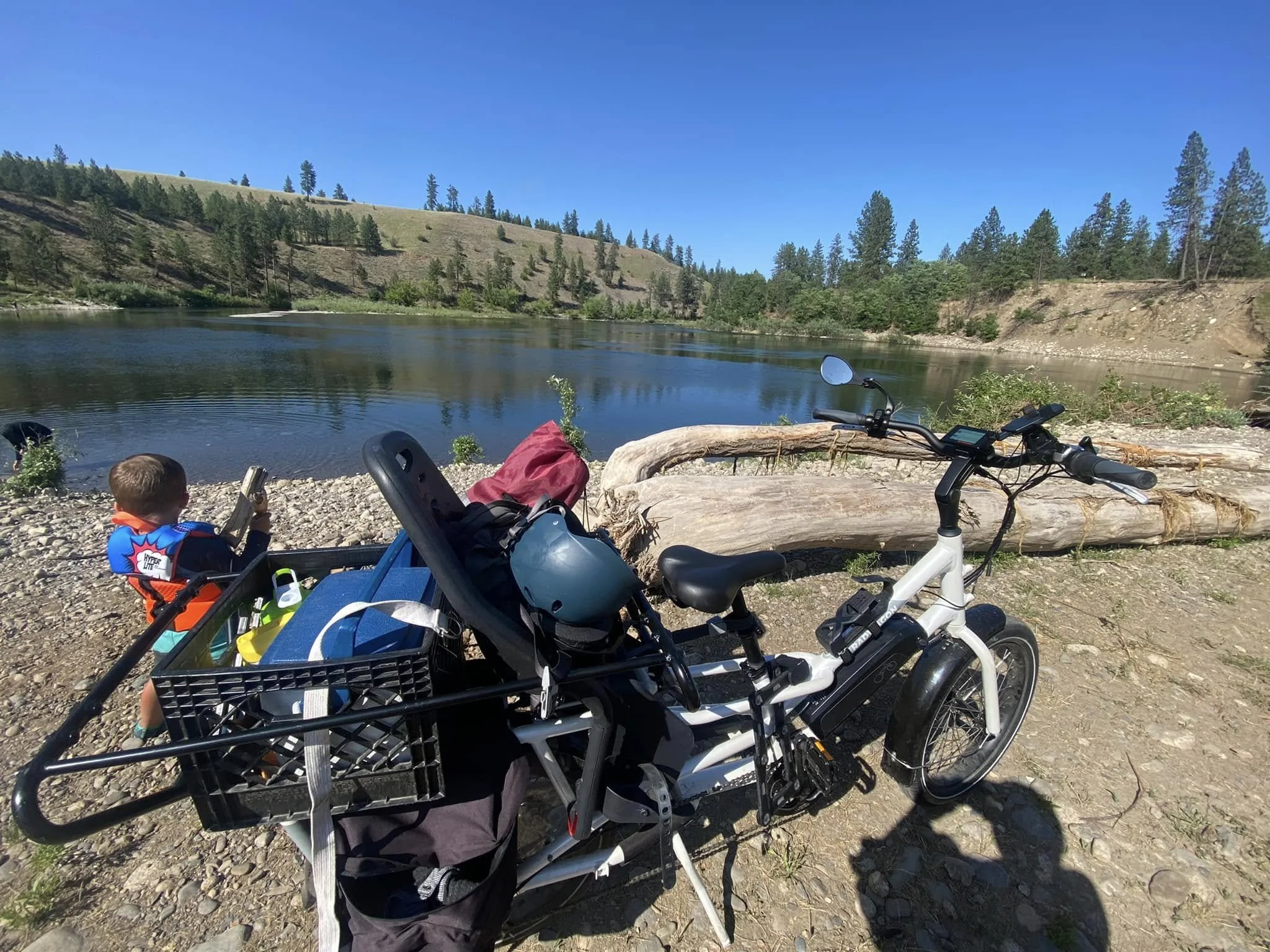 A scenic view of a river with a hillside covered in trees in the background. In the foreground, there are bicycles, a child's helmet, and a child's head visible, indicating outdoor recreational activity near the water.