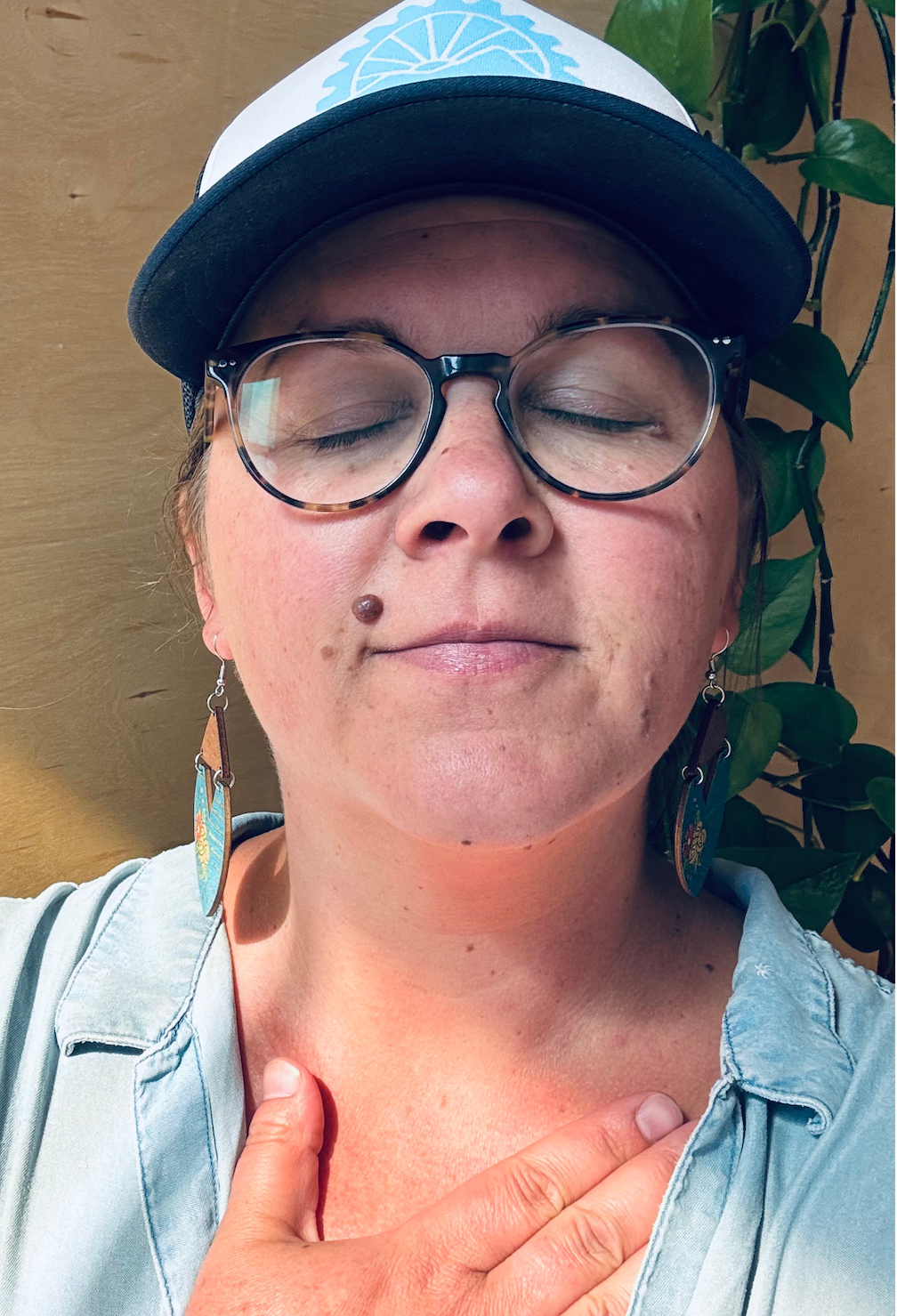 Close-up of a woman wearing glasses, a baseball cap, and earrings, with her eyes closed and hand on her chest, standing against a wooden wall and green plant in the background.