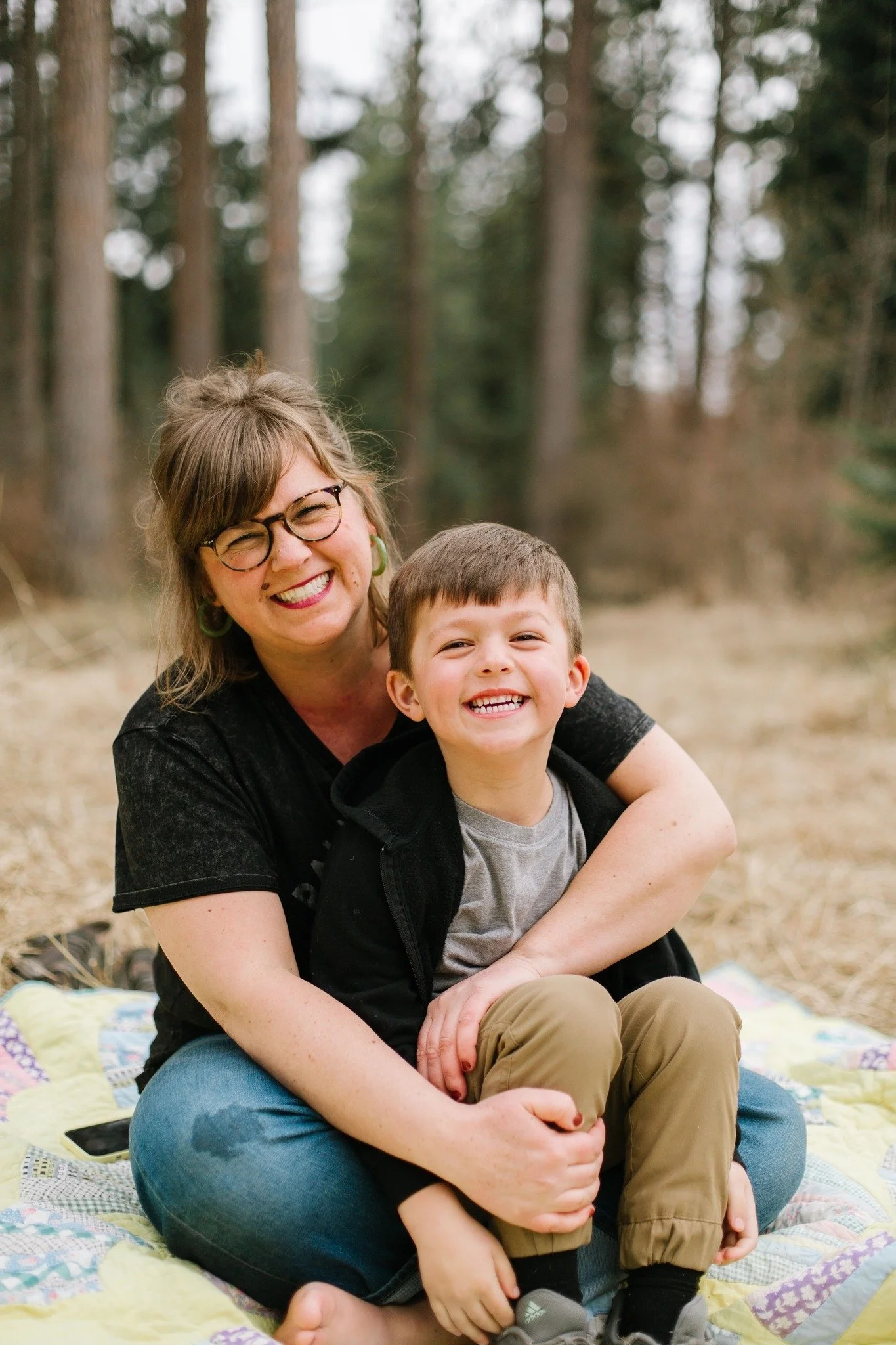 A woman and a young boy sitting on a quilt outdoors, smiling, with trees in the background.