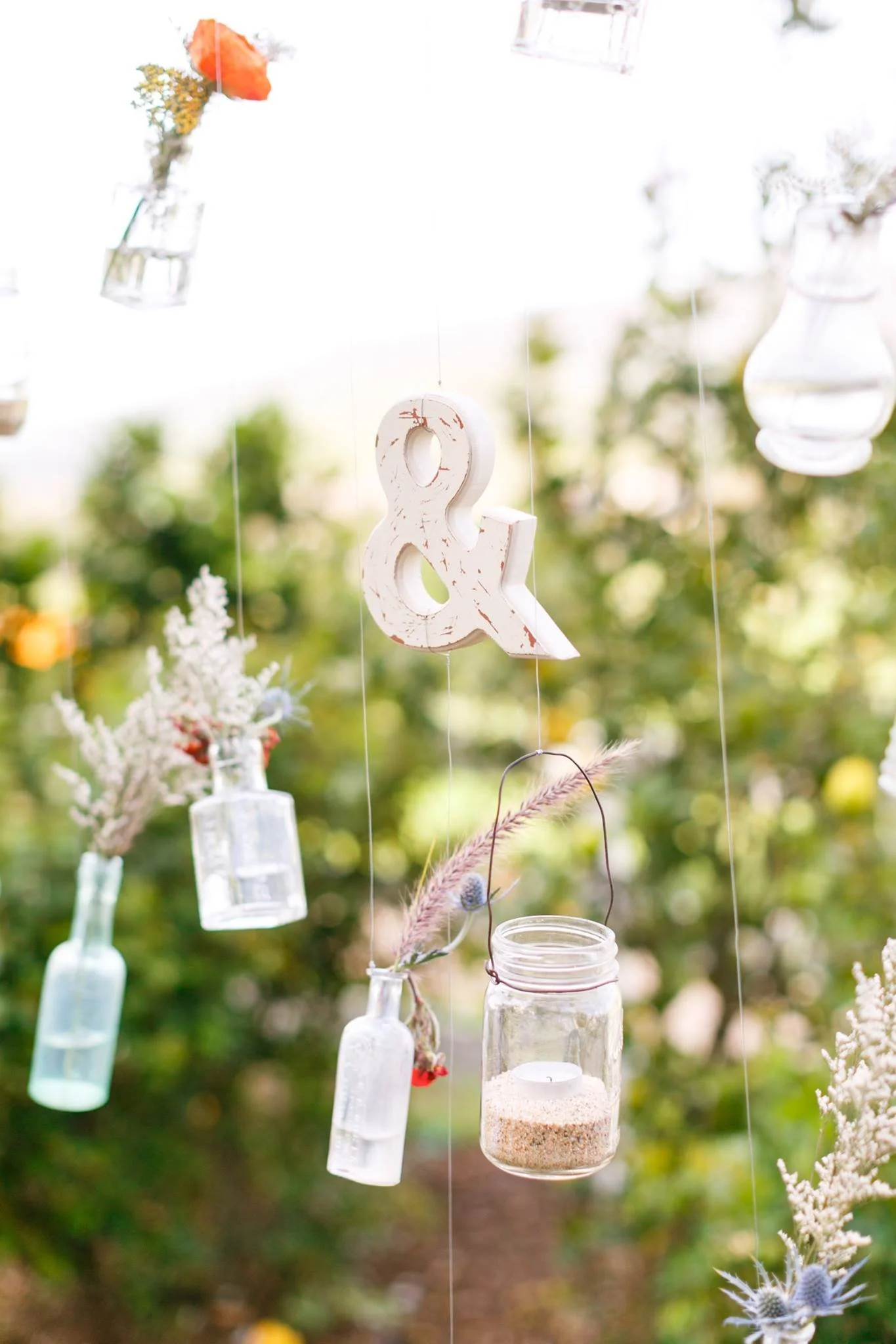 Hanging glass jars and bottles with decorative items such as an ampersand cutout, dried flowers, and feathers, suspended outdoors in a garden setting.