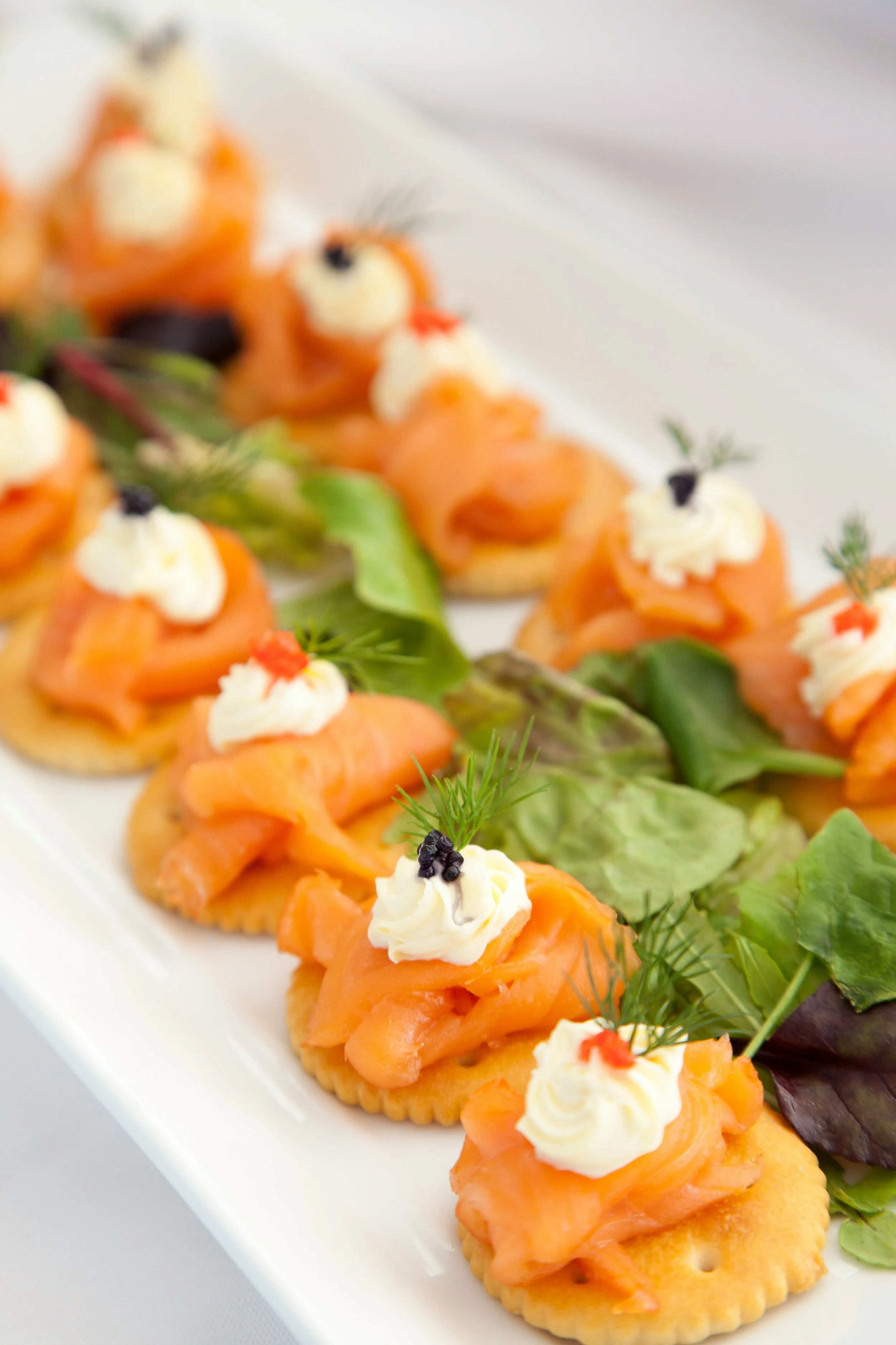 Salmon canapés topped with cream and caviar, garnished with herbs, arranged on a white platter with mixed greens.