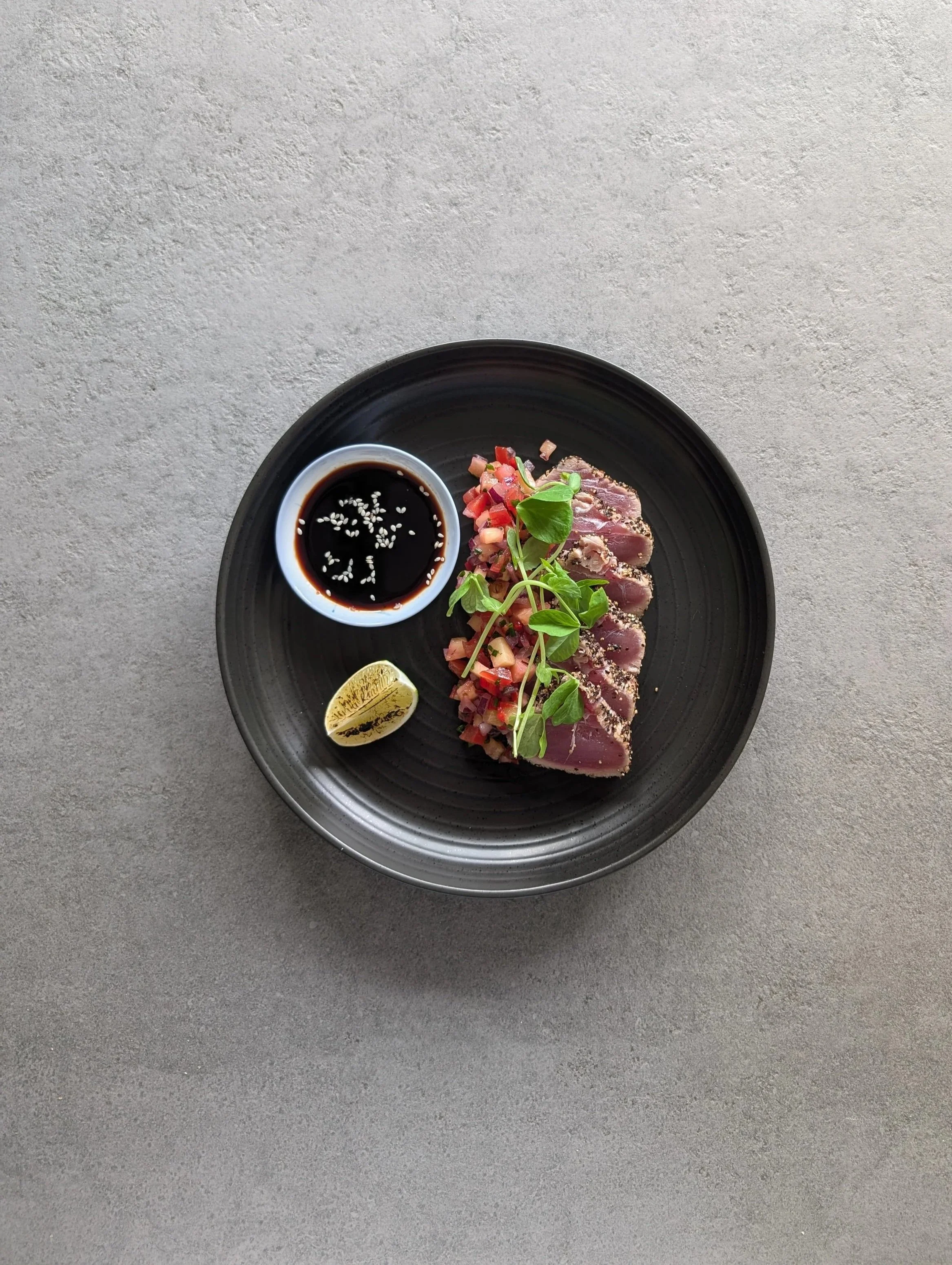 Chef Katie serves seared tuna slices garnished with greens, accompanied by a small dish of soy sauce with soy and ginger dipping sauce and sesame seeds, with a lemon wedge on the side.