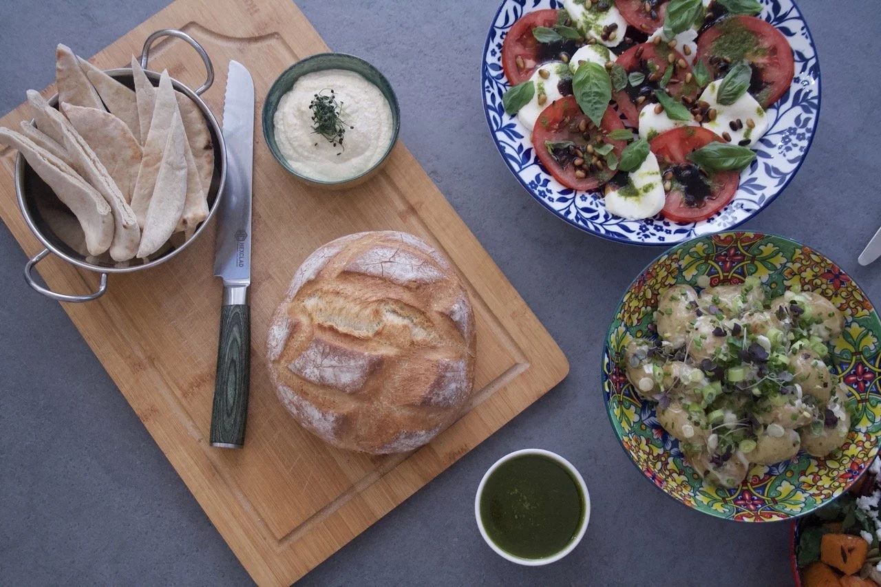 A flat lay of a bread bowl with sliced bread, a bowl of creamy dip garnished with herbs, a small cup of green sauce, a plate of sliced tomato and mozzarella salad with basil, and a plate of potato salad with herbs and microgreens, all on a gray surface with a wooden cutting board.