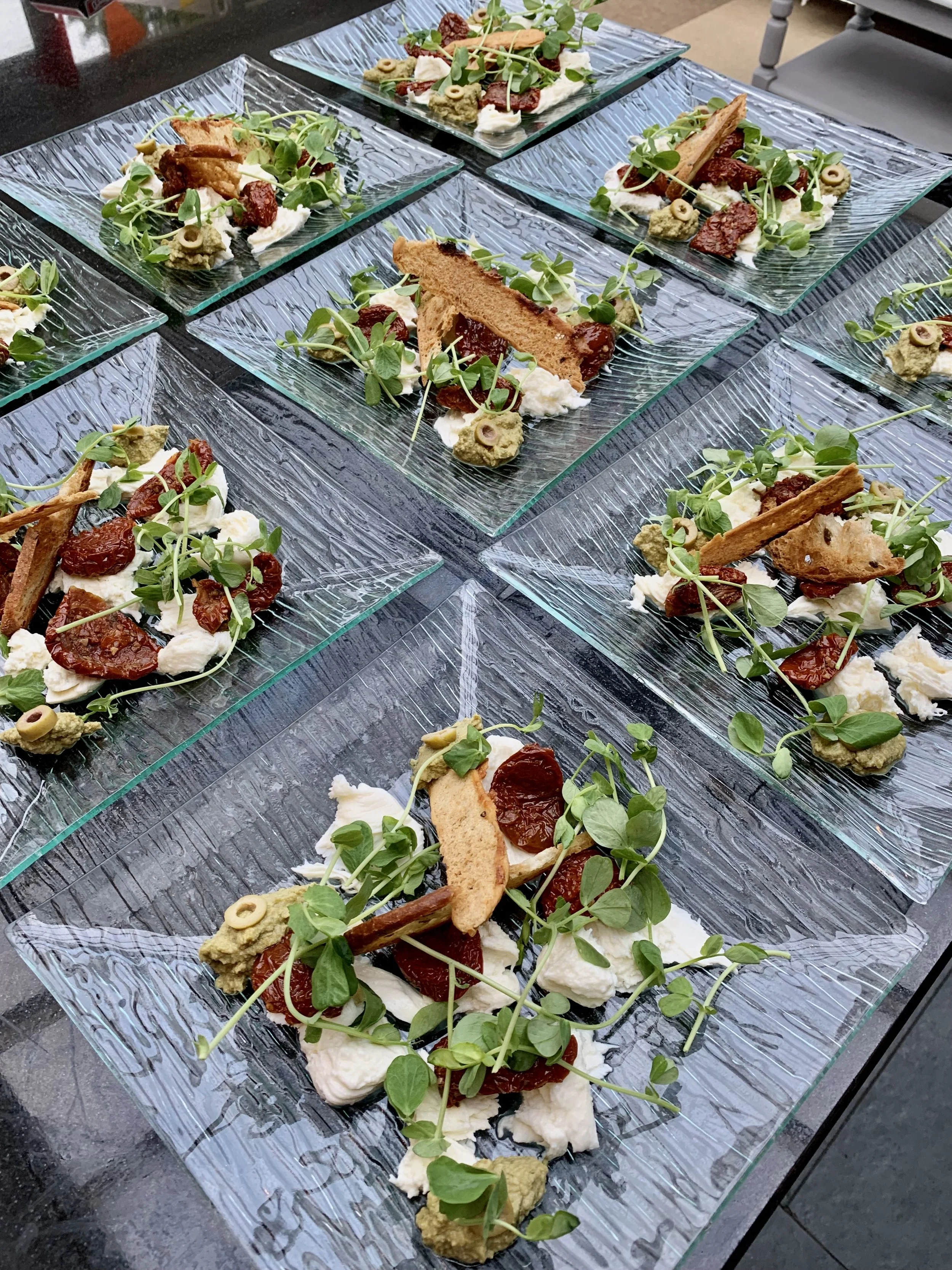 Multiple servings of gourmet antipasto on decorative glass plates, garnished with greens such as microgreens, and topped with various cured meats, cheeses, and crackers, arranged on a dark table.