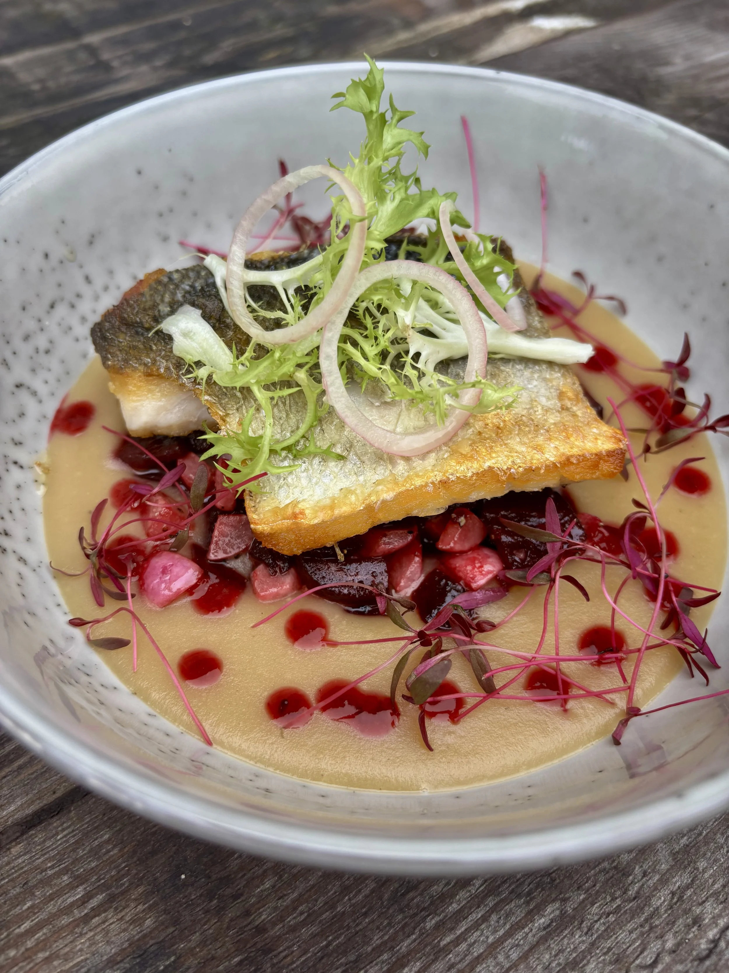 A plated dish featuring seared fish fillet with crispy skin, topped with mixed greens and drizzled with sauces and edible microgreens, served in a white bowl on a wooden table.
