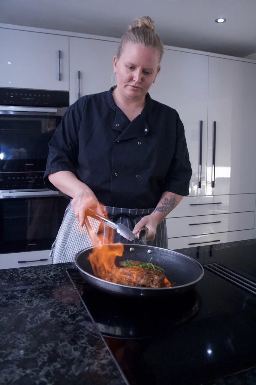 Chef Katie in a black chef's coat is cooking steak in a frying pan on a stovetop, with flames rising from the pan.