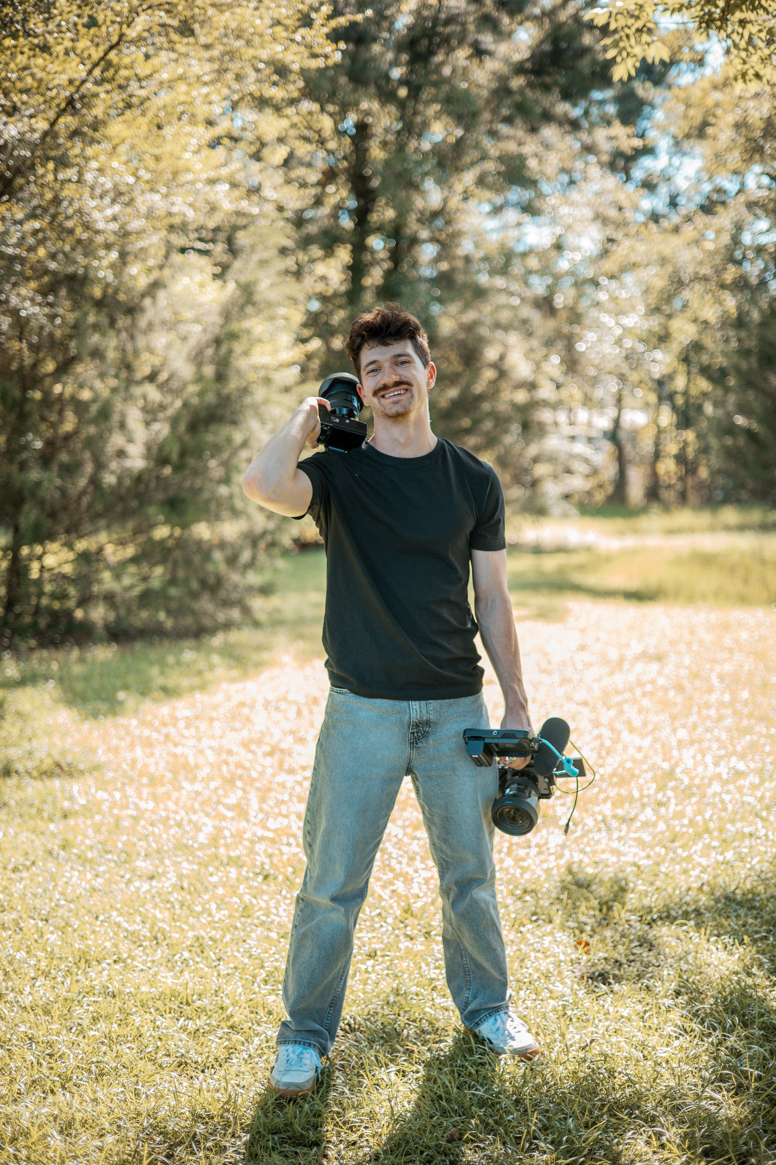 A young man with curly hair and a mustache smiling outdoors in a park with trees. He is holding a professional camera in his right hand and a drone with a remote control in his left hand, standing on a grassy area during sunlight.