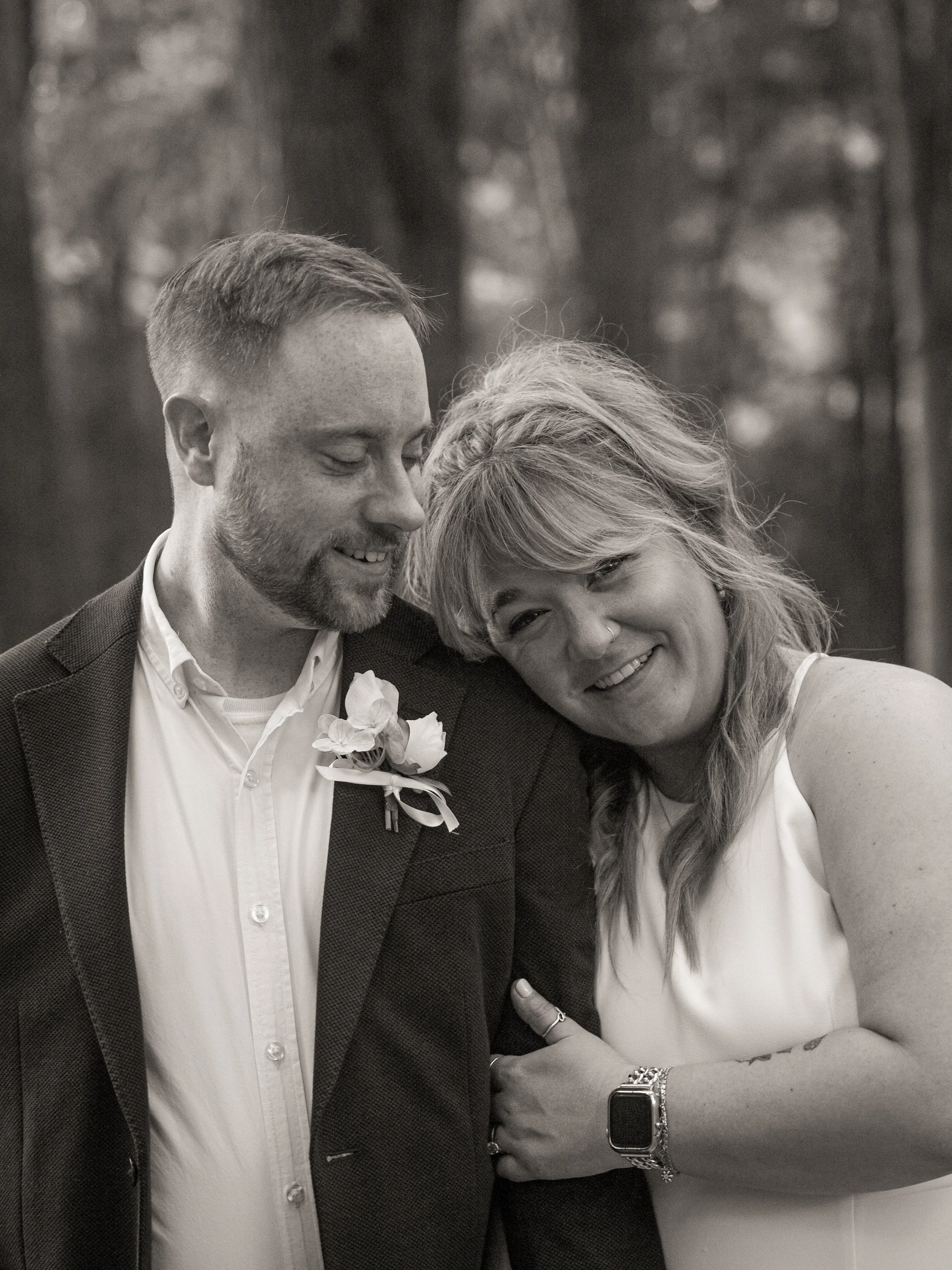 A black and white photo of a man and woman smiling and sharing a moment together, with the woman resting her head on the man's shoulder. The man is wearing a suit with a boutonniere, and the woman is wearing a sleeveless dress with a watch and bracelet on her wrist. They seem happy and affectionate.
