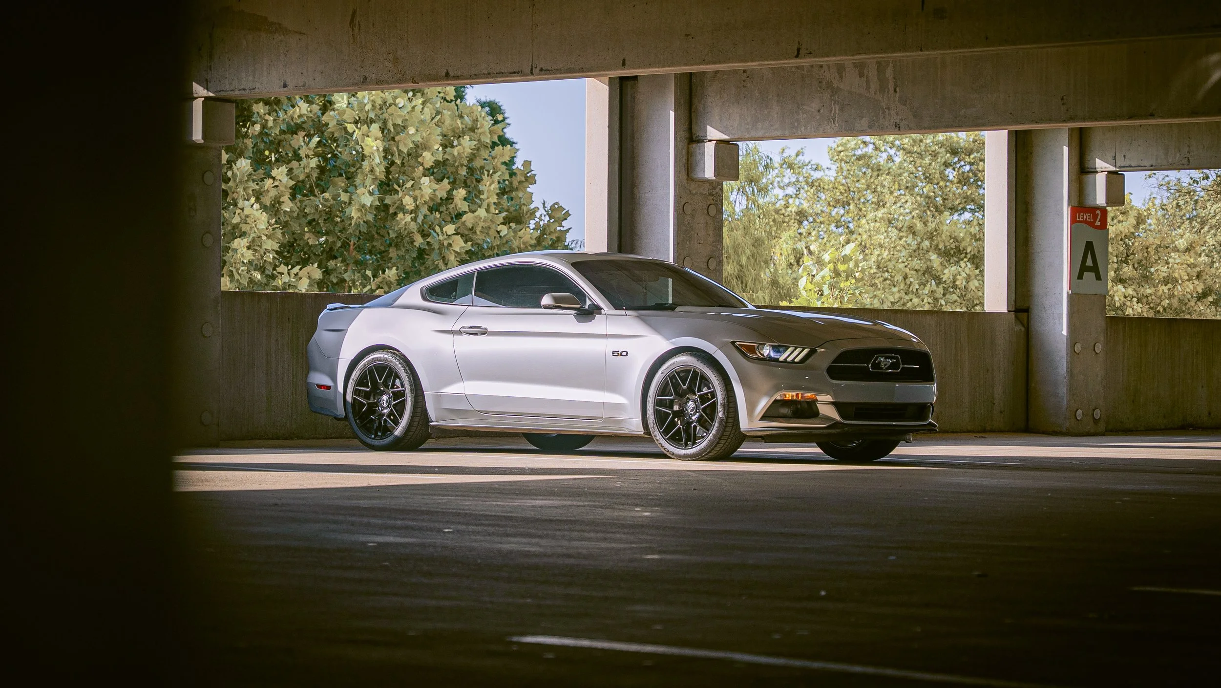 A silver Ford Mustang parked in an outdoor parking garage. The photo is taken from a low angle, with the car framed by the concrete structure of the garage and some trees in the background.
