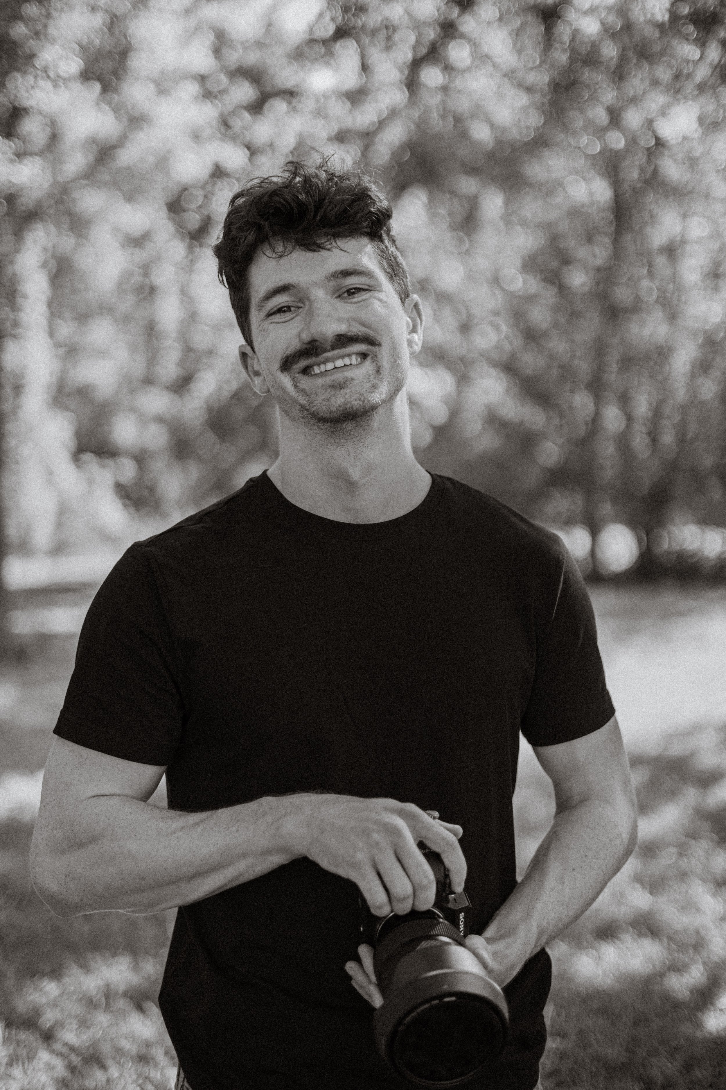A smiling man with curly dark hair and a mustache, wearing a black t-shirt, holding a camera with a large lens. He is outdoors with blurred trees in the background.