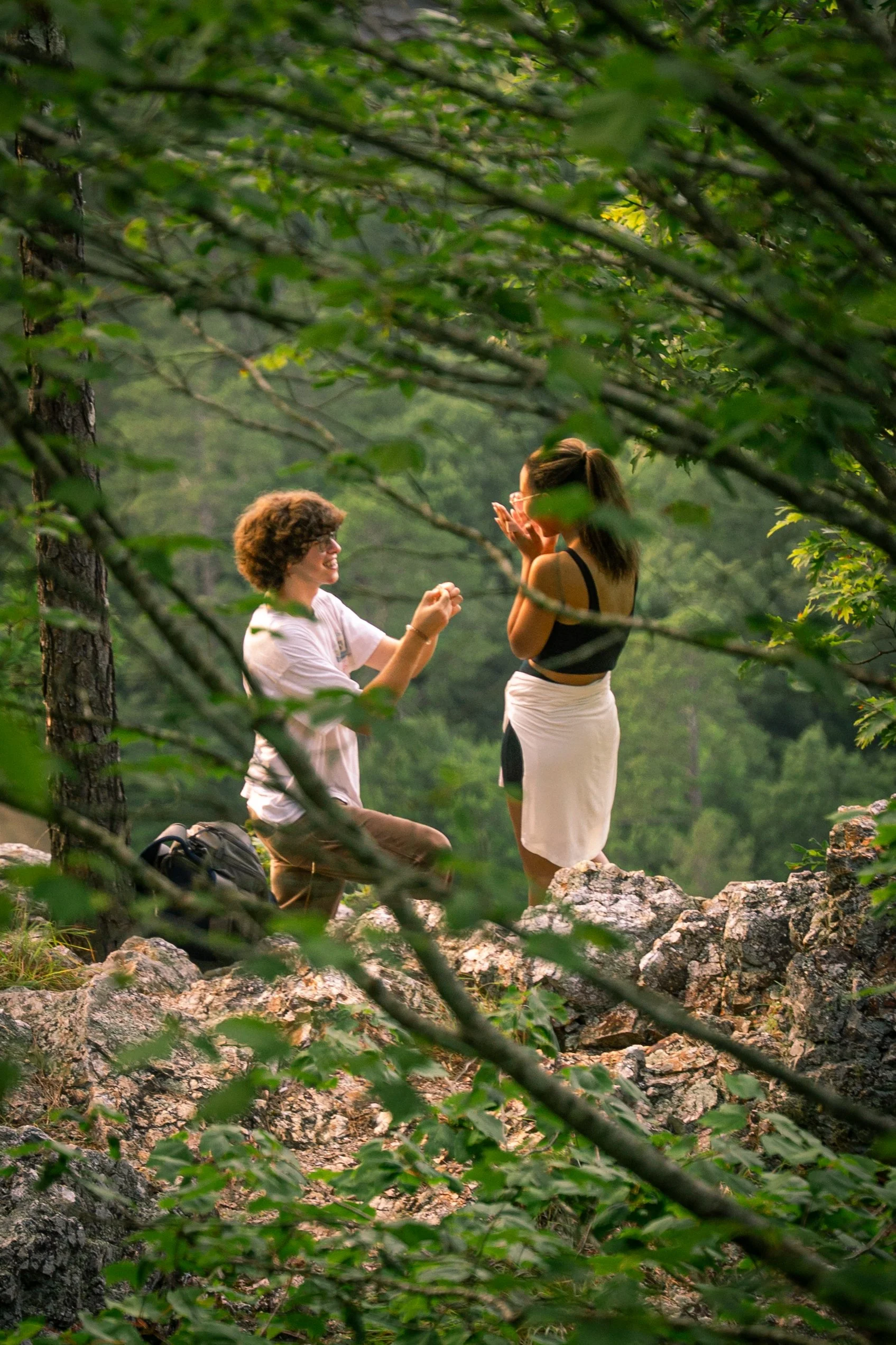 A man and his girlfriend are communicating in a lush forest, with one kneeling and the other standing on a rocky terrain, surrounded by dense green leaves and trees.