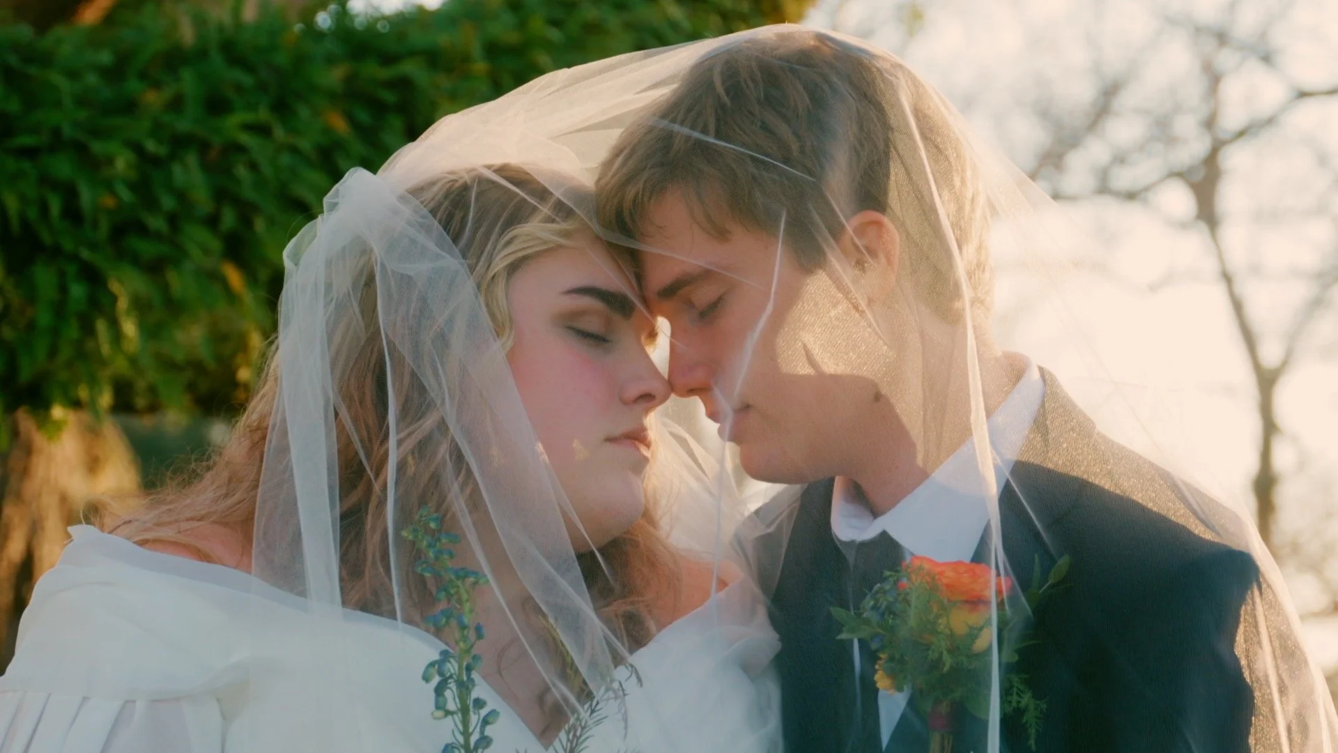 A bride and groom with closed eyes, touching foreheads, sharing a tender moment under a veil during their outdoor wedding ceremony, with trees and sunlight in the background.