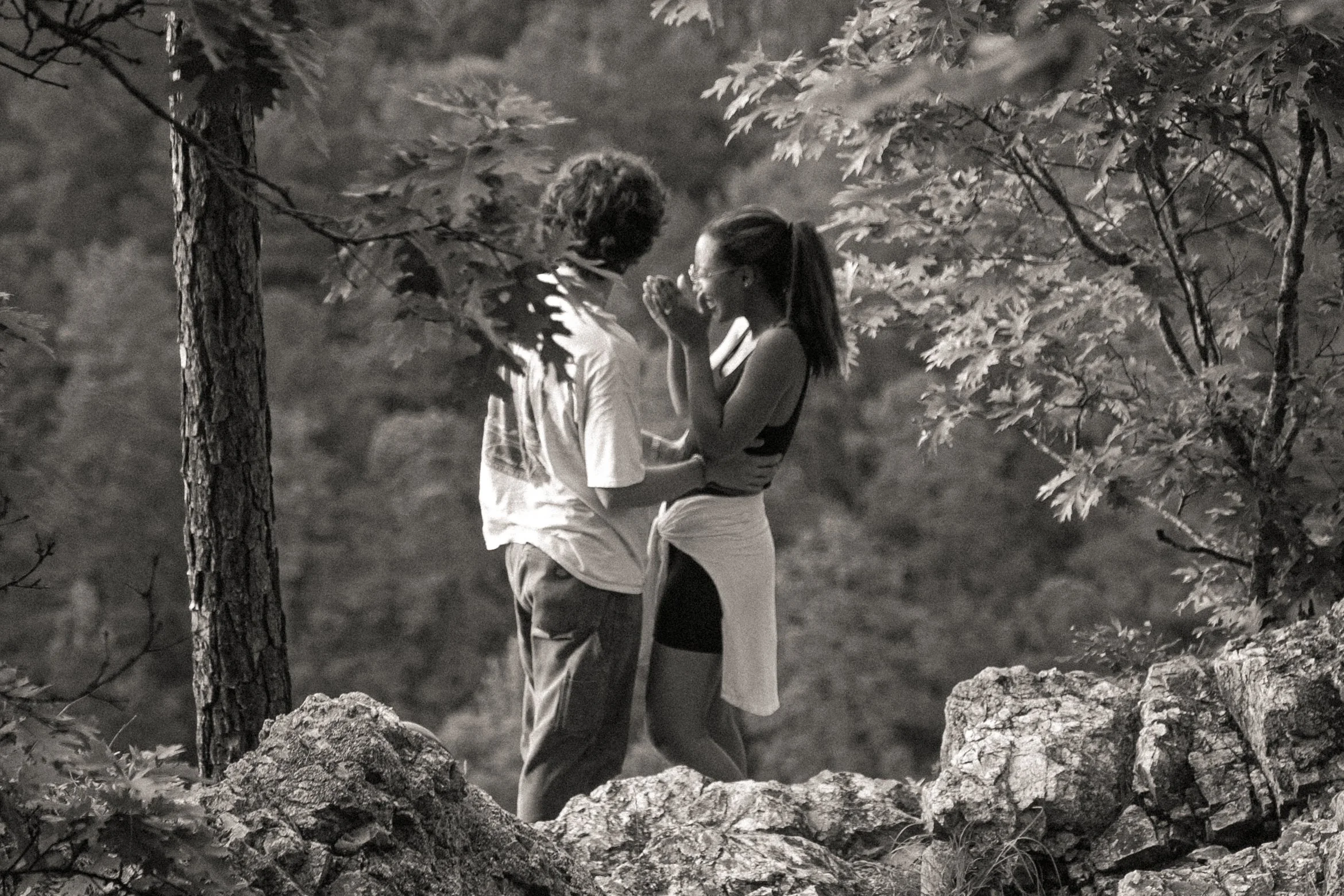Two people, a man and a woman, standing outdoors among trees and rocks, engaging in a close, intimate moment, possibly sharing a kiss or whispering, with a blurred natural background.