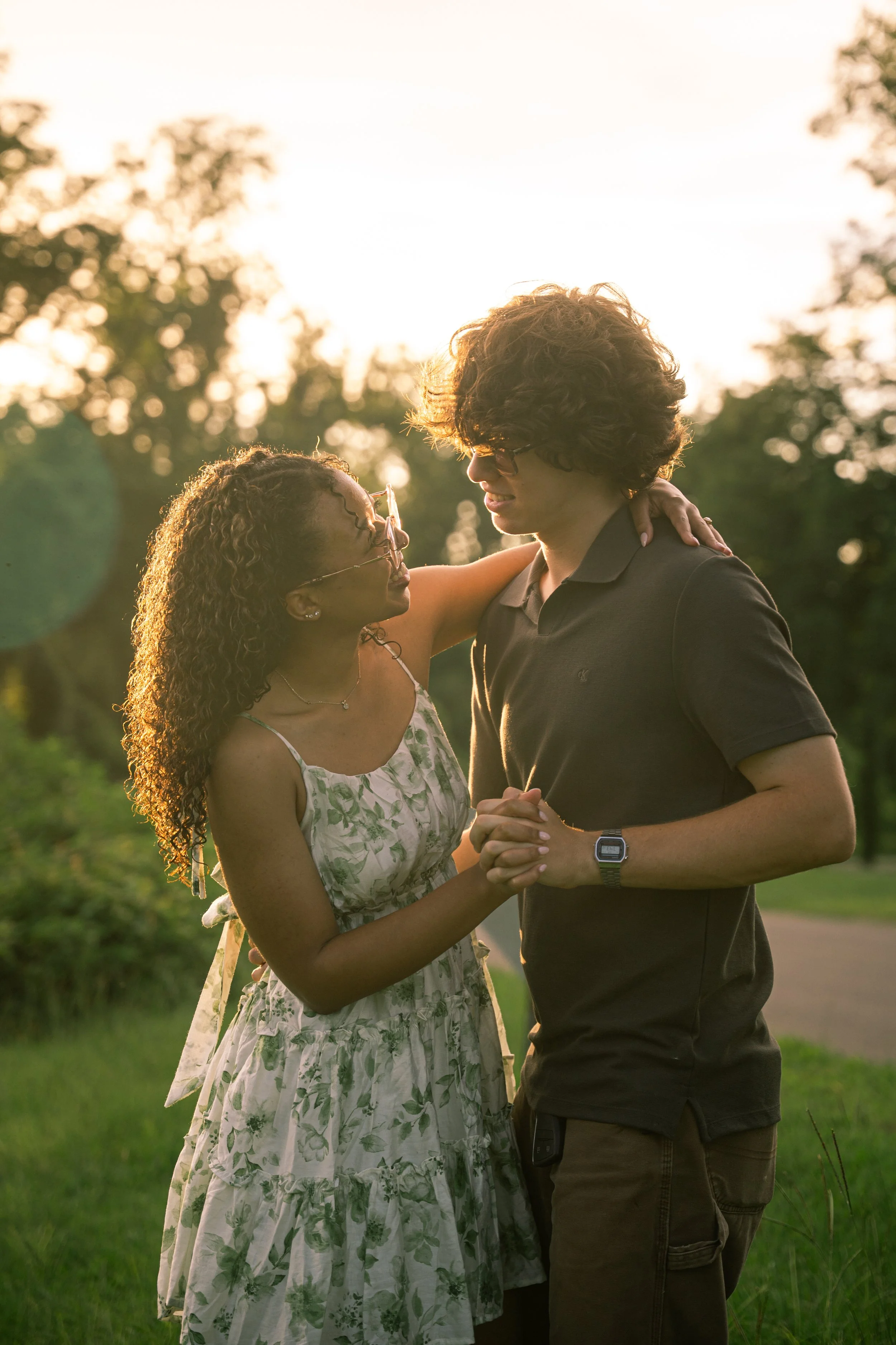 A woman and a man share a tender moment outdoors during sunset, holding hands and gazing into each other's eyes, with trees in the background.