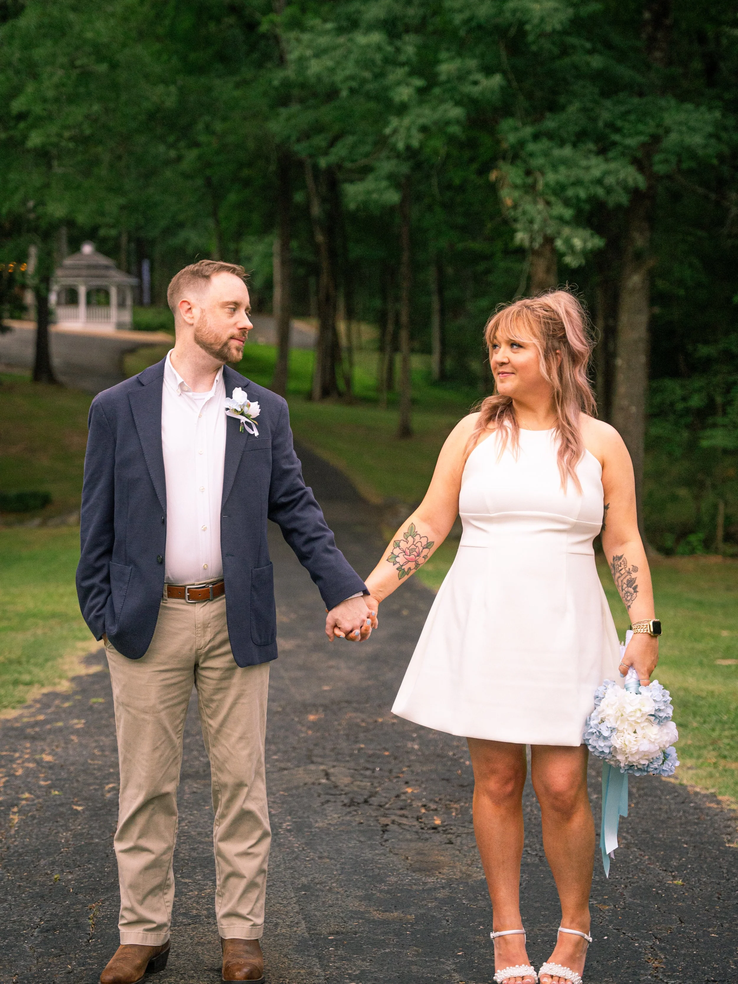 A couple holding hands outdoors on a paved path in a wooded area, dressed in wedding attire, with the woman holding a bouquet and wearing high heels and the man in a blazer and khaki pants.