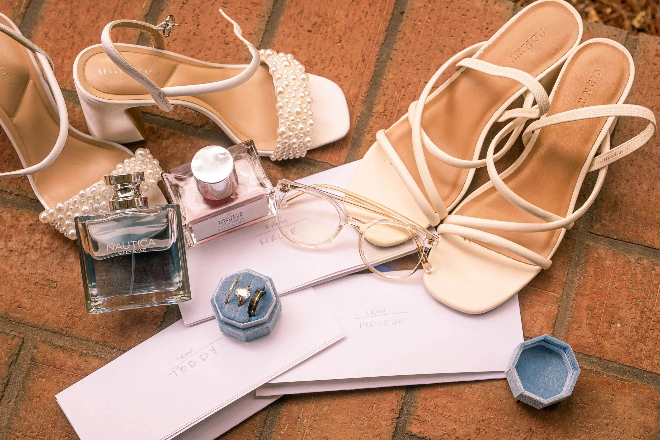 Pair of white high-heeled shoes with ankle straps and pearl embellishments, a pair of white sandals, perfume bottles, jewelry, and sunglasses arranged on a white paper and brick floor.
