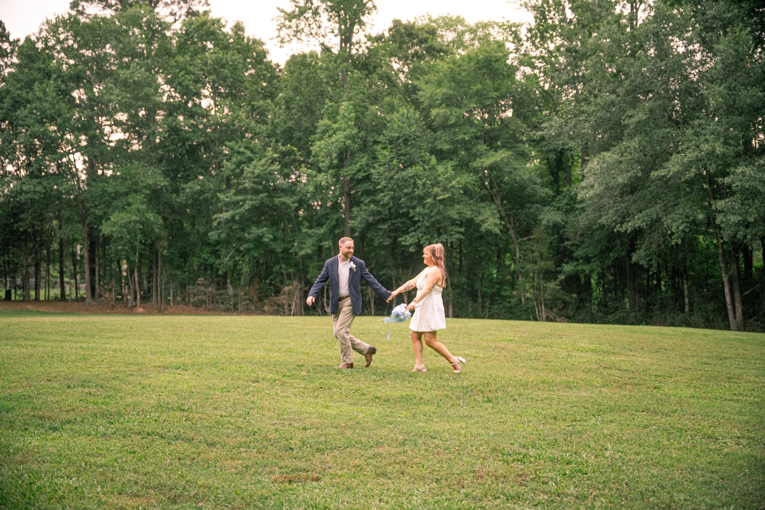 A couple dancing happily outdoors on a green field with a background of tall trees and an overcast sky. The man is wearing a dark blazer, light pants, and a boutonniere, while the woman is wearing a white dress and holding a bouquet.