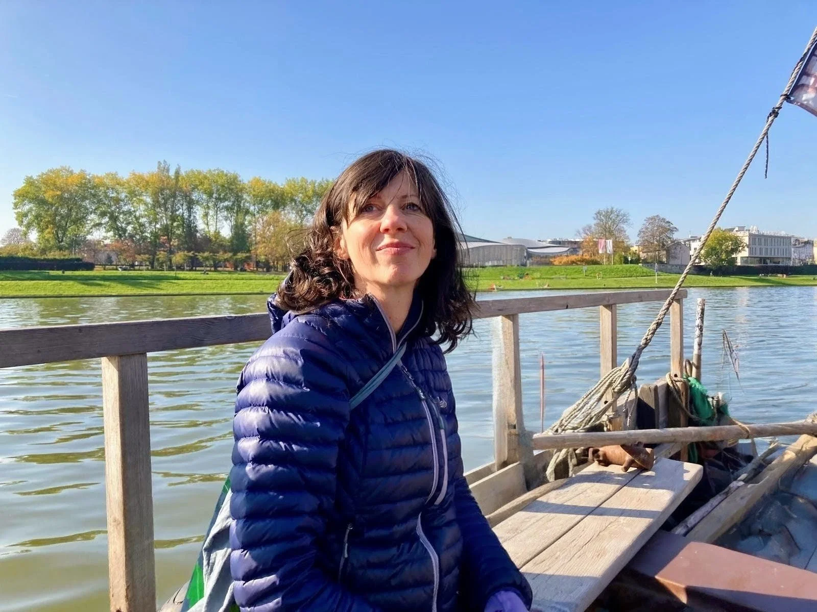 A woman with dark hair wearing a blue puffer jacket sitting on a wooden boat near a body of water with trees and buildings in the background on a sunny day.