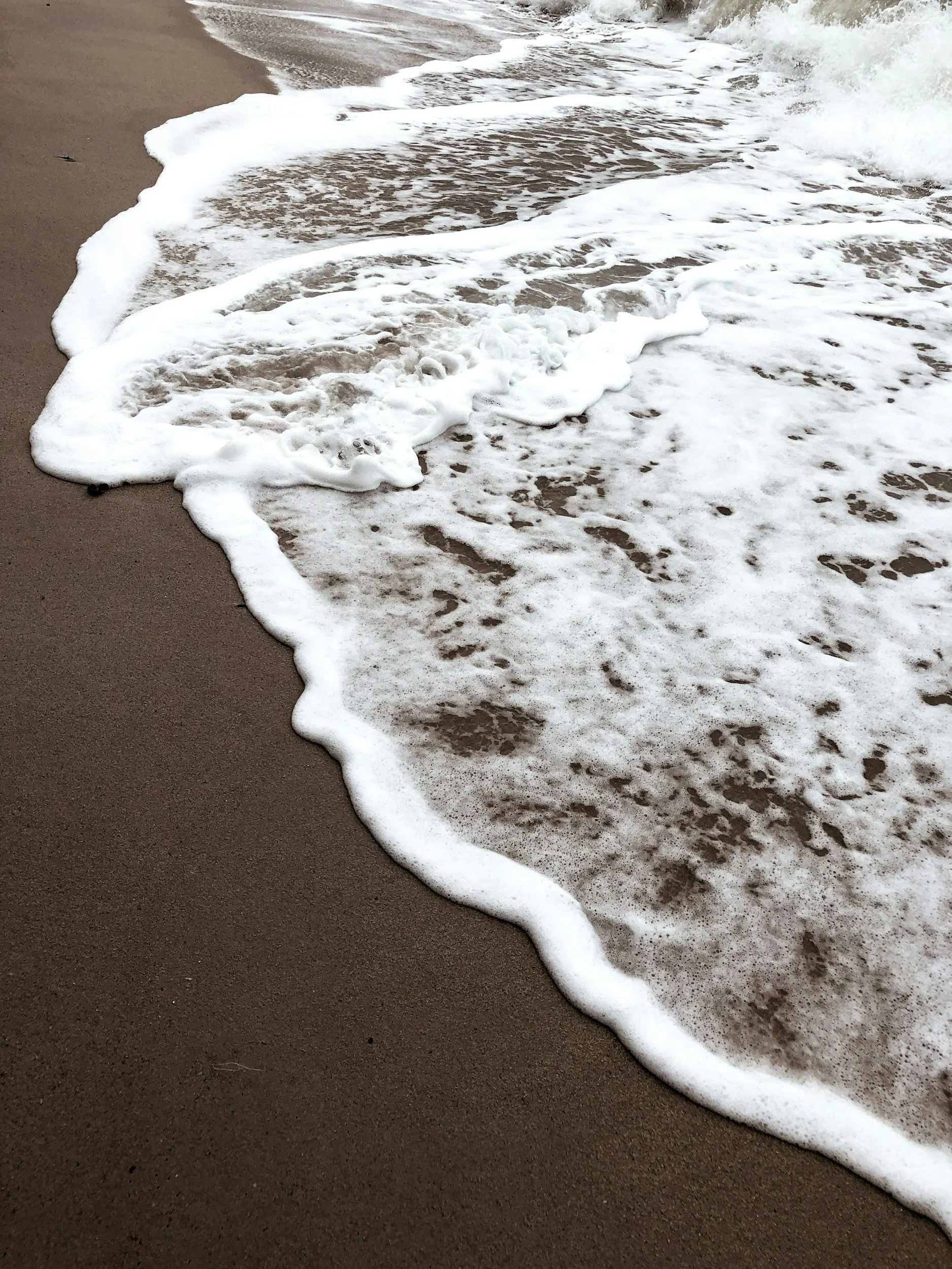 Close-up of ocean waves gently washing onto a sandy beach, forming white foam on the dark brown sand.