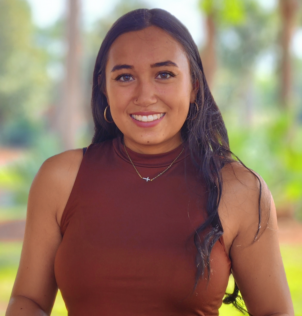 A young woman with long dark hair, wearing a sleeveless brown top, gold hoop earrings, a necklace with a cross, and smiling outdoors with a blurred background of trees and greenery.