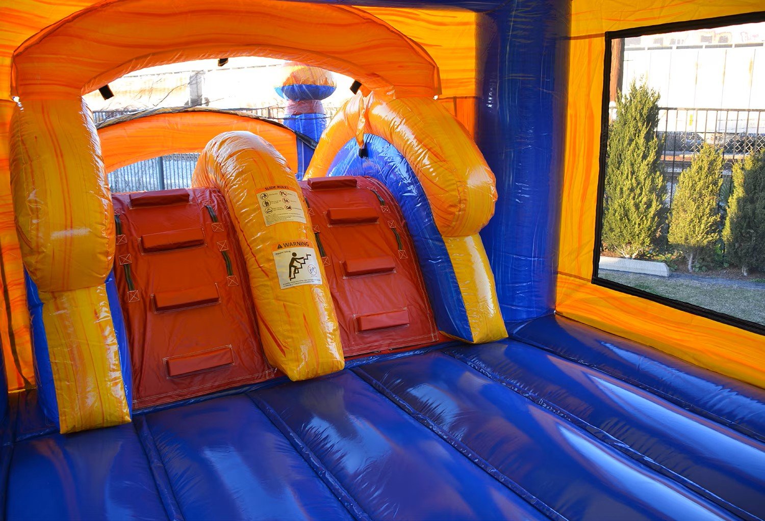 Interior of a colorful inflatable bounce house with a slide and climbing area, orange, yellow, and blue sections, with a safety warning label on the slide.